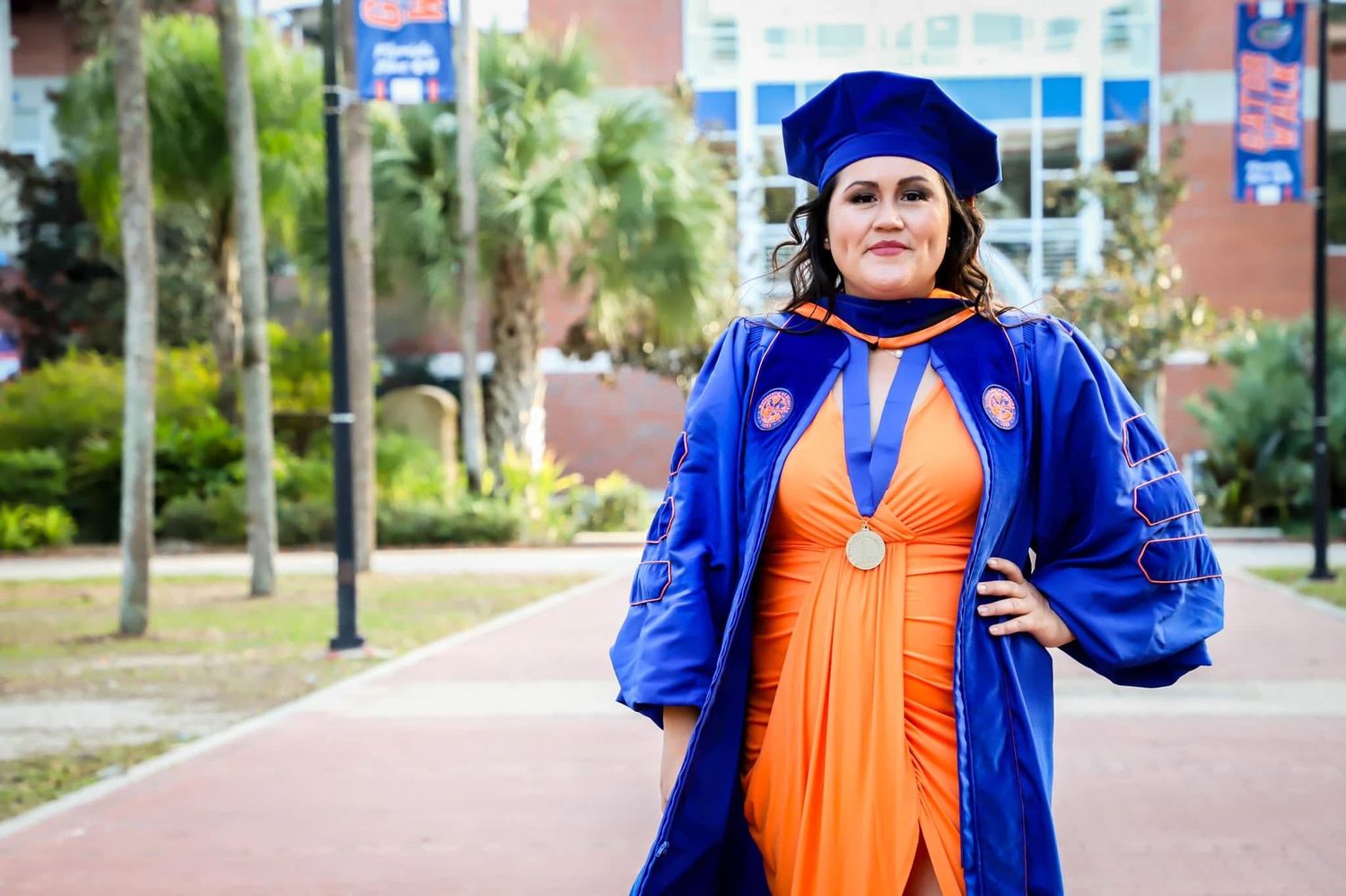 Doctoral graduation portrait of a woman in a blue gown and orange dress posing on a university campus walkway in Gainesville, Florida.