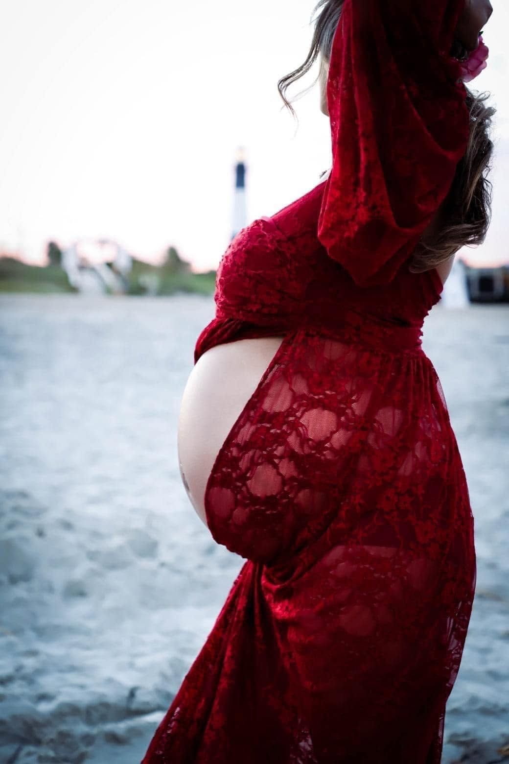 A pregnant woman in a red lace hooded gown cradles her baby bump on a sandy beach near a lighthouse in Georgia