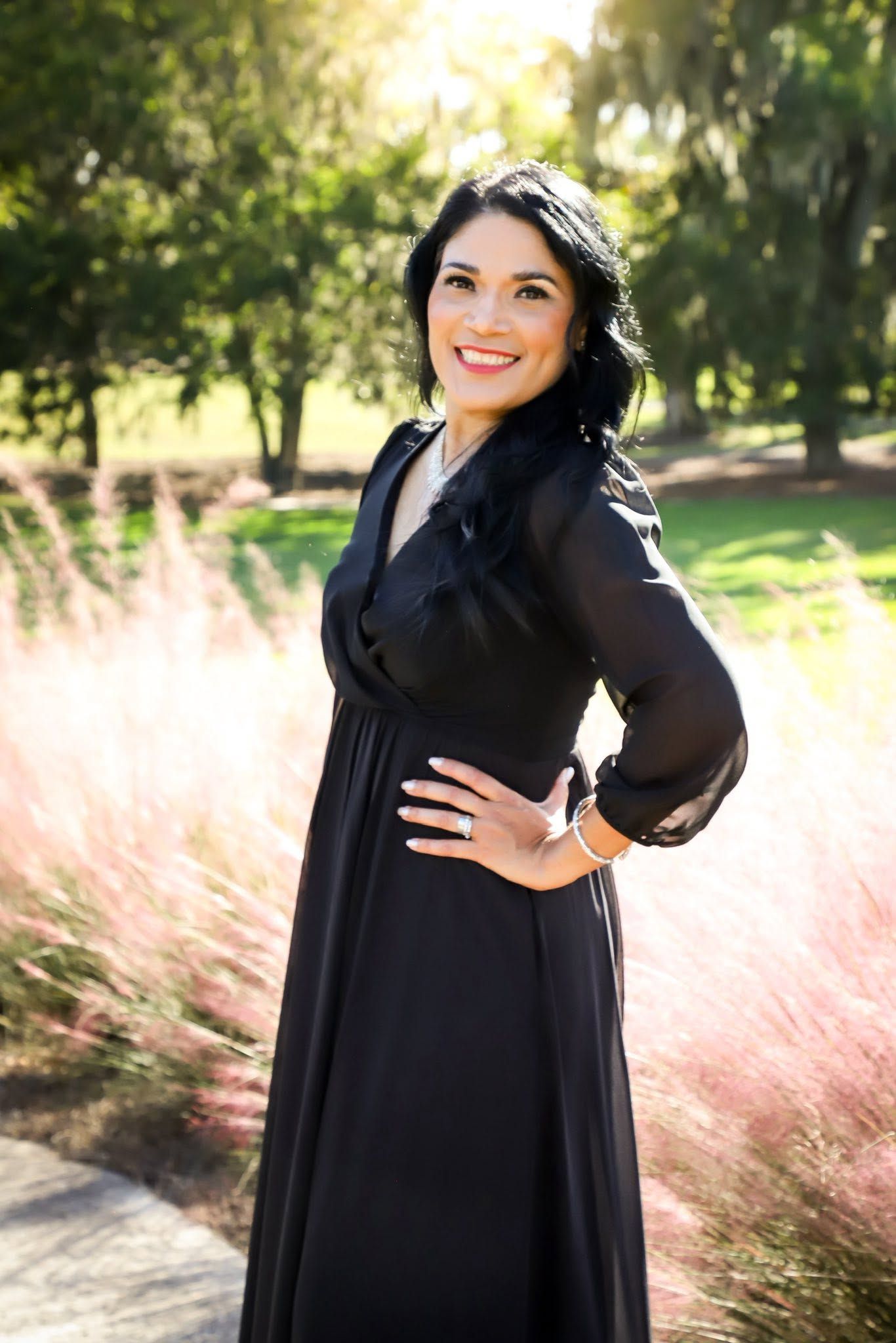 Woman in black maxi dress smiling outdoors among pink ornamental grasses in a sunlit park.