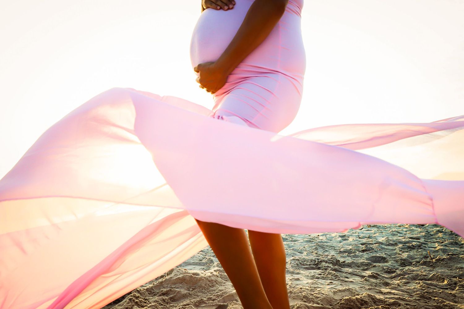 Maternity Photography Session with Pregnant woman in flowing pink dress holding baby bump on sandy beach at sunset in Jacksonville Florida