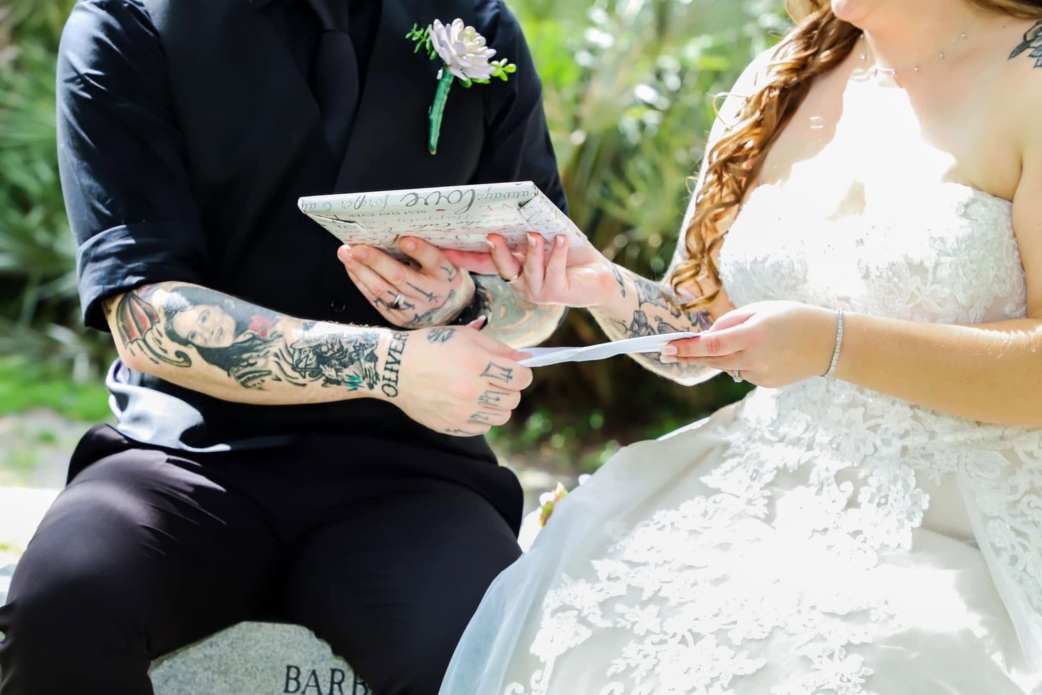 Tattooed groom and bride in lace dress exchange vows documents while seated during outdoor wedding ceremony.