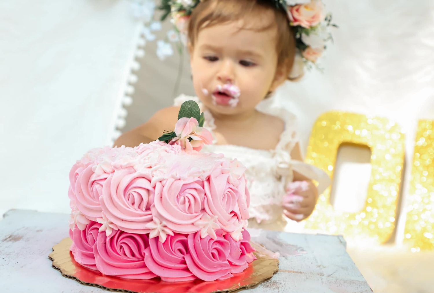A baby girl with a floral crown reaches into a pink ombre rose-swirl birthday cake in Gainesville, Florida.