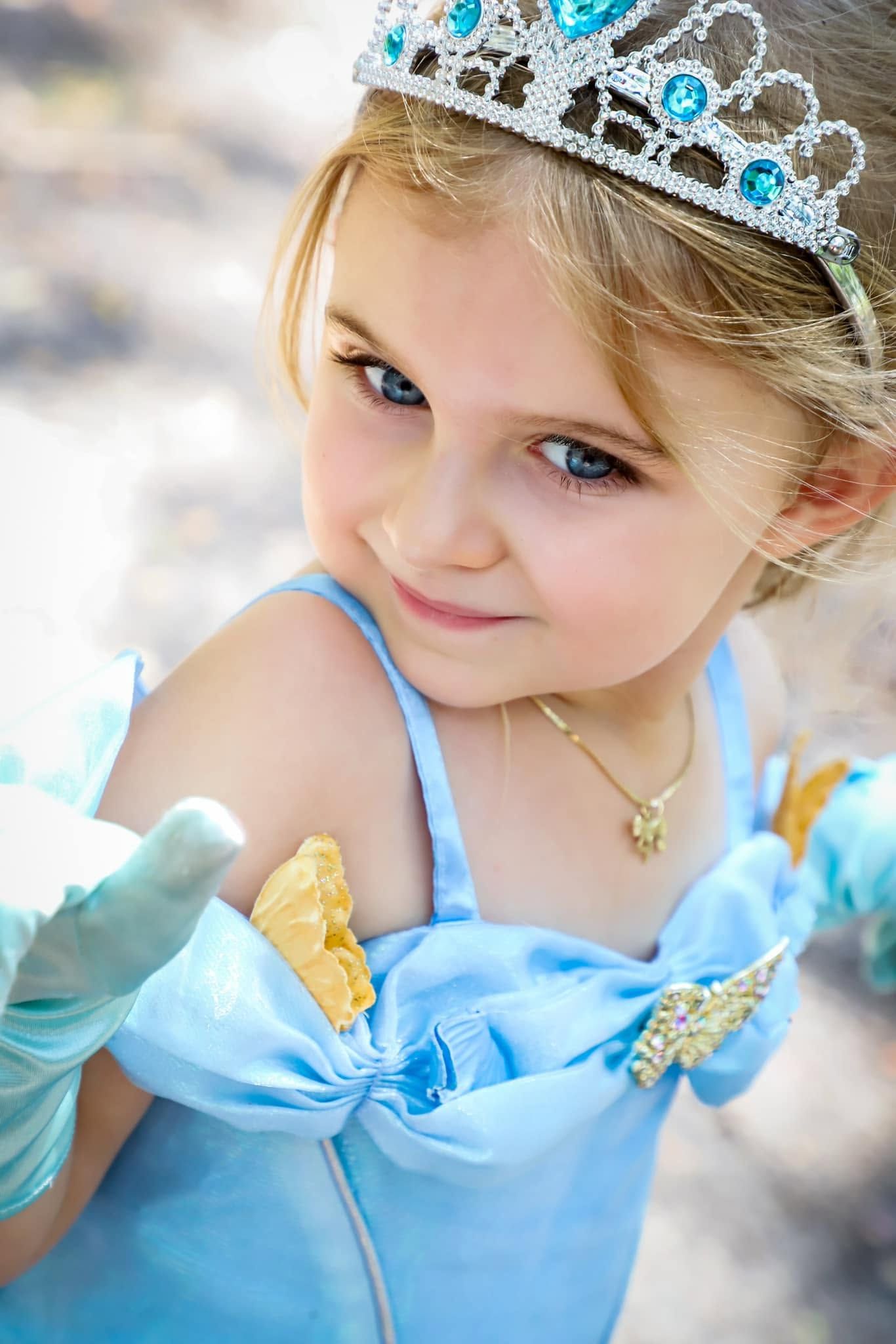 A young girl dressed as a princess in a blue Cinderella costume with a tiara and gloves smiles sweetly.