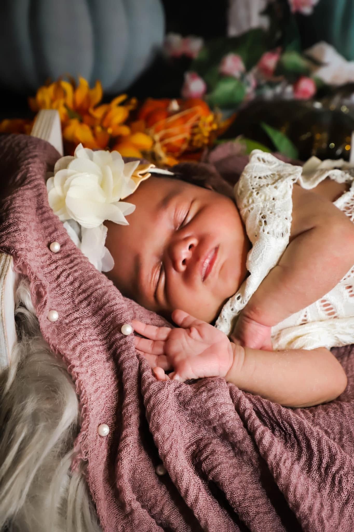 Peaceful sleeping newborn baby girl in white lace outfit with flower headband on pink blanket with autumn decor in Georgia home studio.