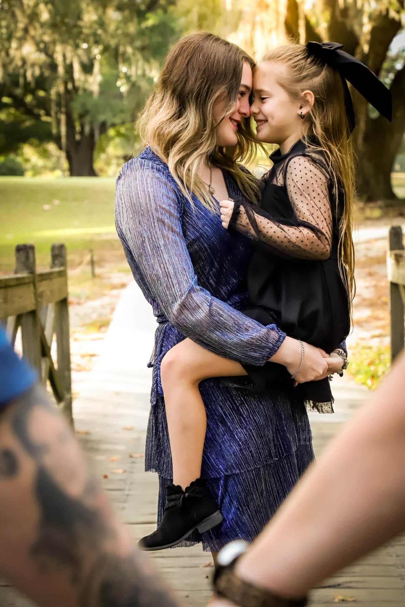 Mother in blue dress holding young daughter in black outfit on a wooden bridge outdoors.