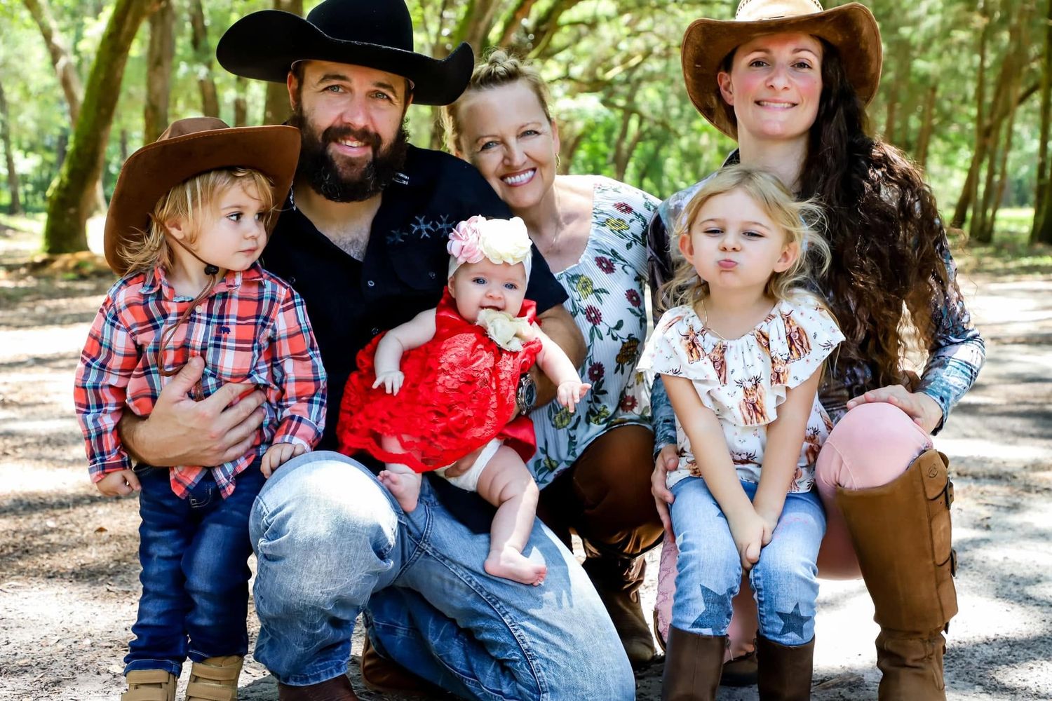 Happy family with three young children in cowboy hats posing outdoors under shady trees.