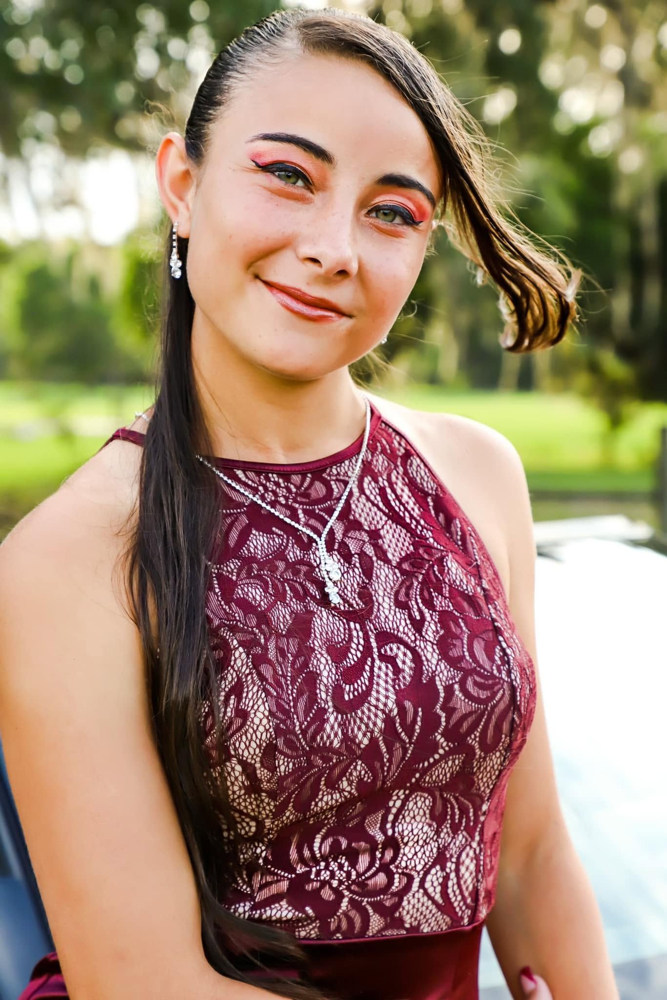 Promo Photography. Smiling teenage girl in burgundy lace dress with bold red eye makeup at outdoor prom photo session.