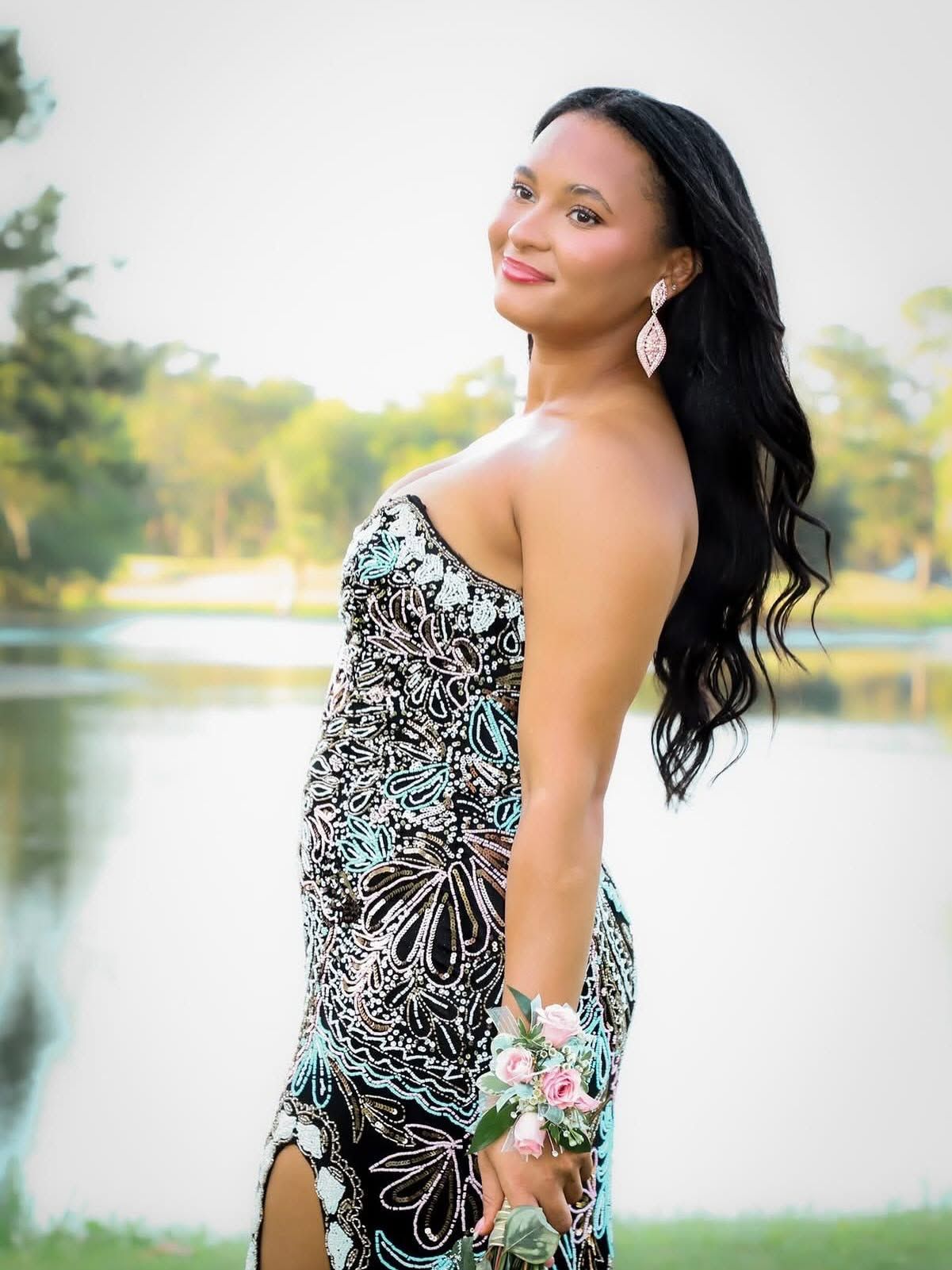 Young woman in elegant floral prom dress with wrist corsage posing near a lake at dusk.