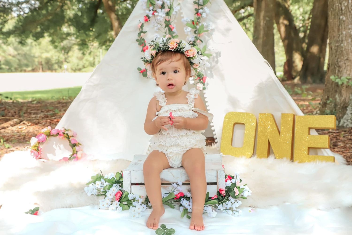 Cake smash photography session featuring a one-year-old girl in a white romper sitting beside a “ONE” sign and floral teepee her first
