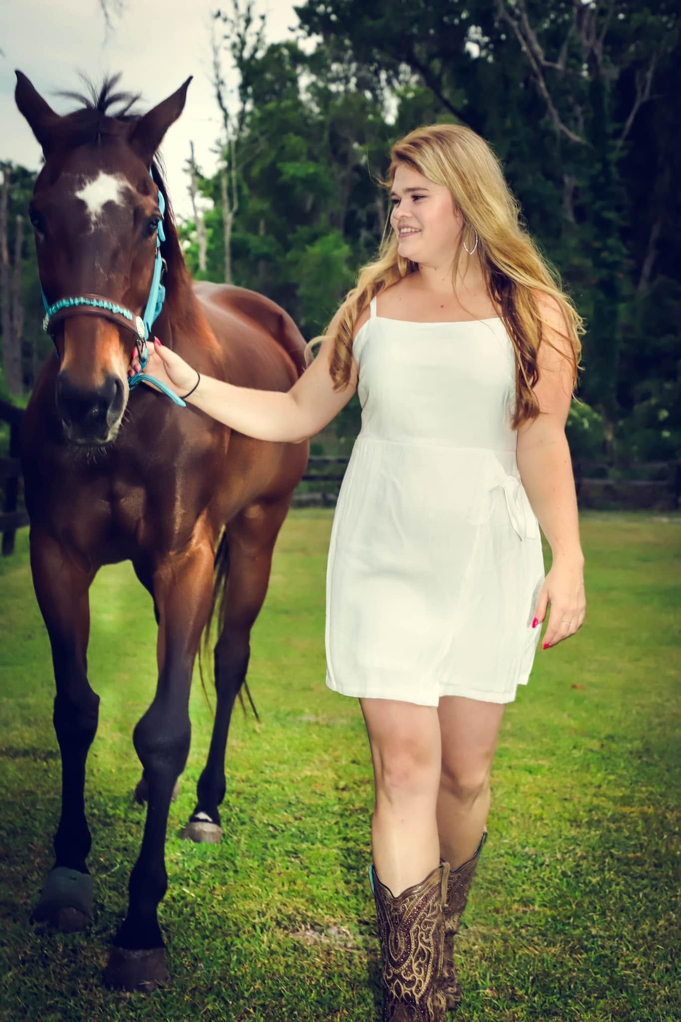 Smiling blonde teenage girl in white dress and cowboy boots walking with brown horse on green grass in Ocala Florida