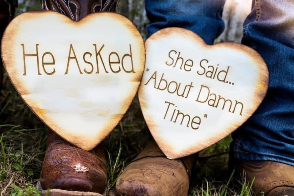Engagement photography session and couple holds two wooden signs reading He Asked and She Said About Damn Time while wearing cowboy boots.