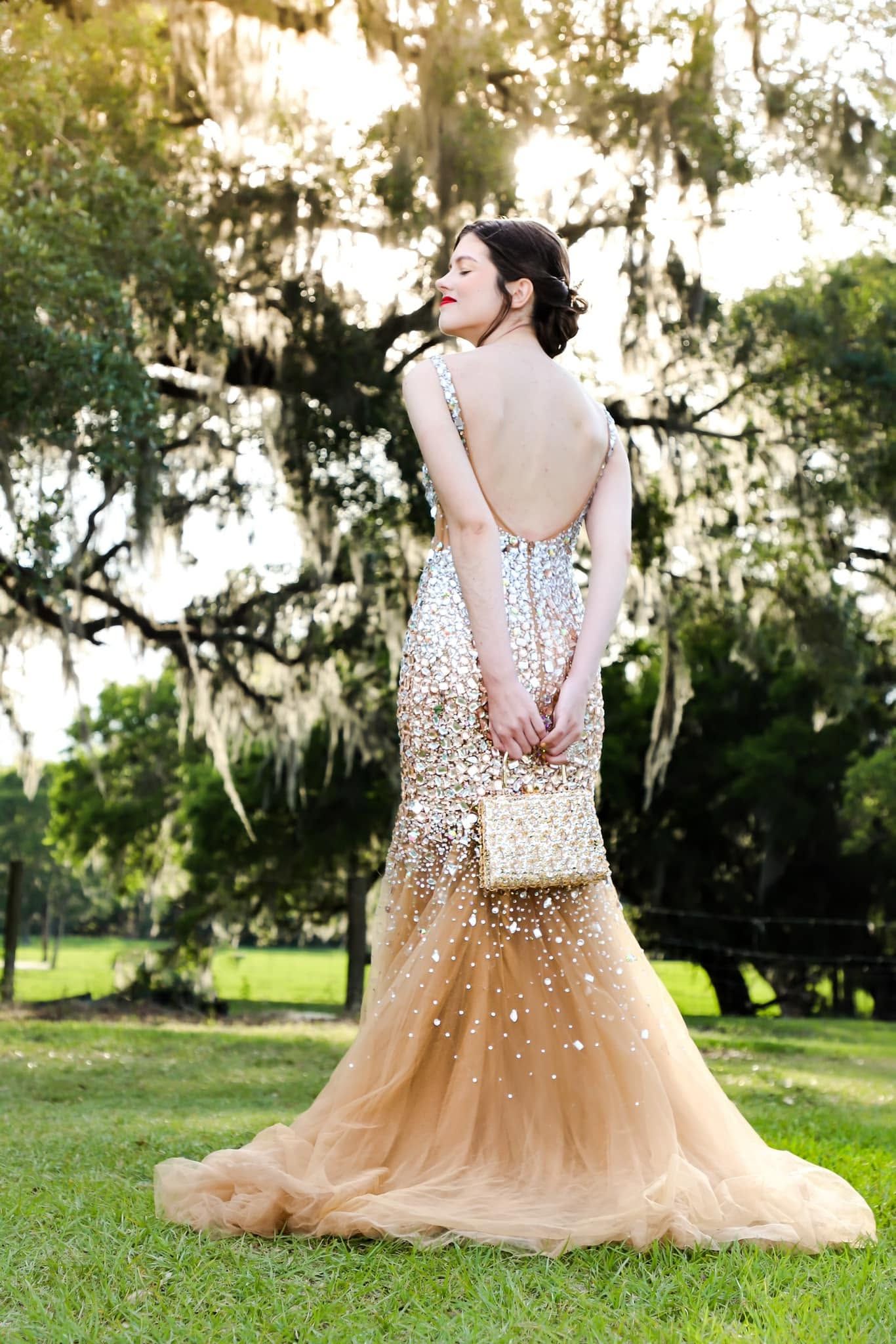 A woman in a stunning backless gold sequined mermaid gown poses outdoors under moss-draped trees at golden hour.