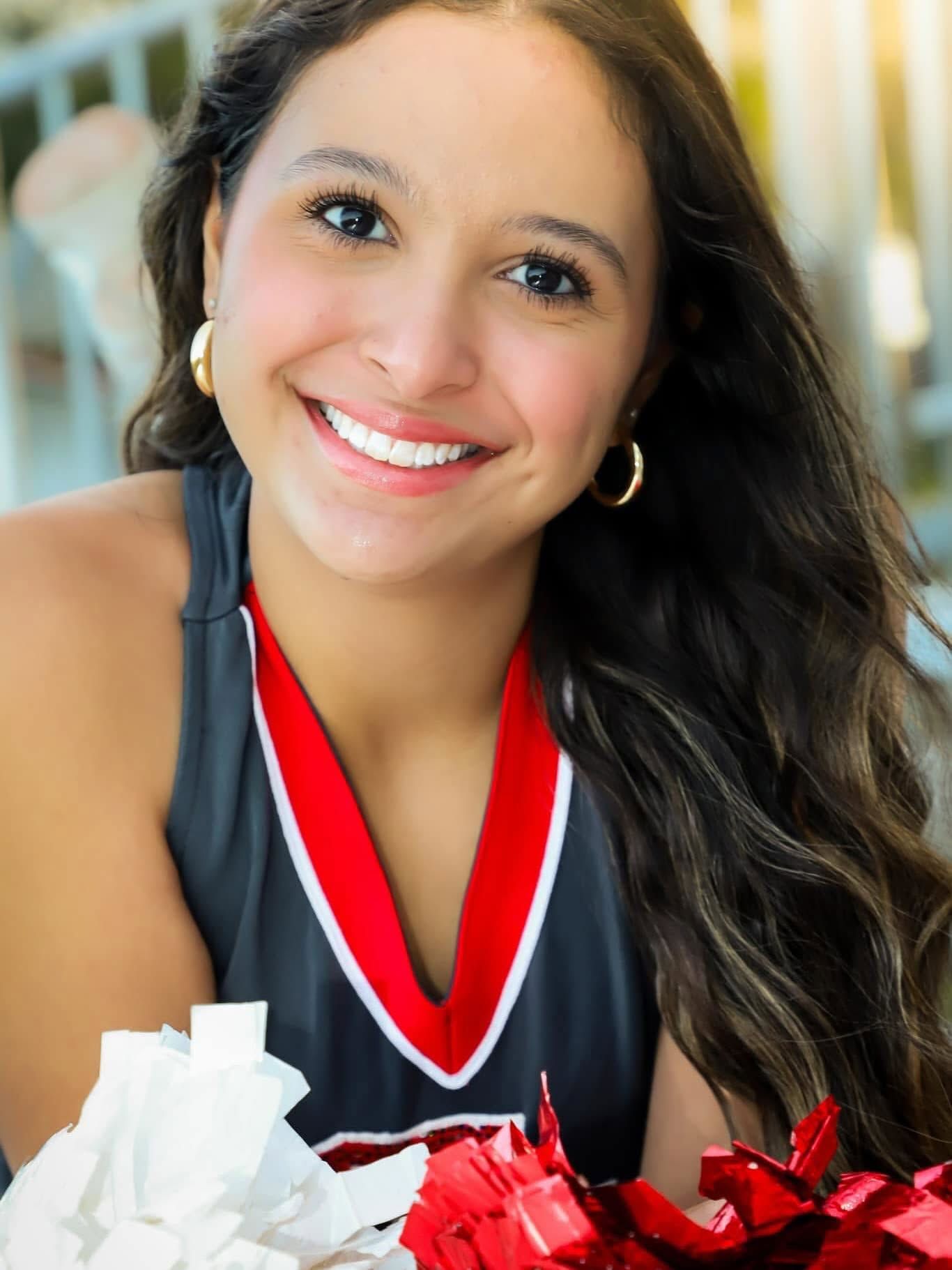 Smiling cheerleader in uniform holding red and white pom poms with hoop earrings.