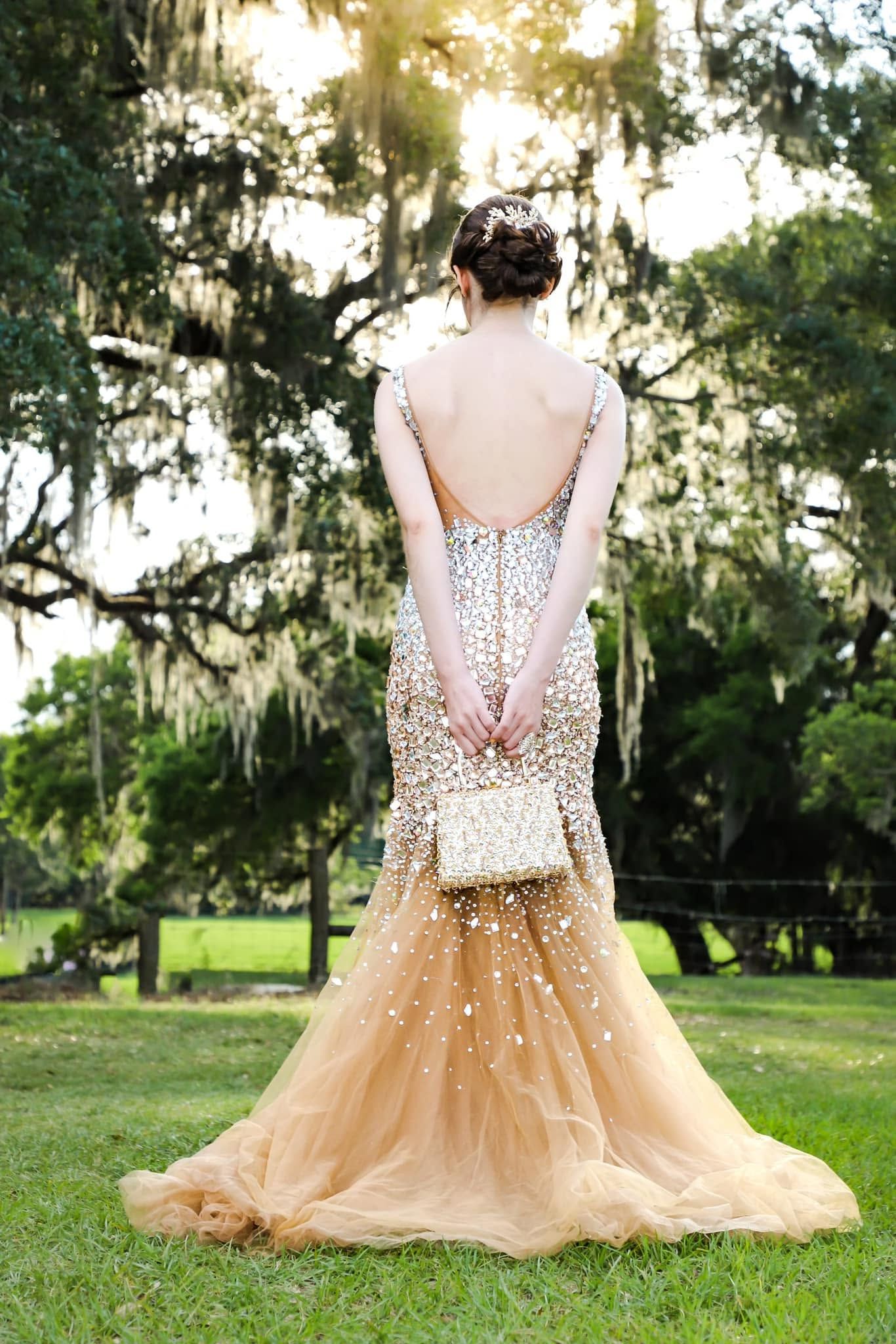 Wedding portrait of an elegant woman in a gold beaded mermaid gown holding a clutch beneath Spanish moss trees in Orlando, Florida.