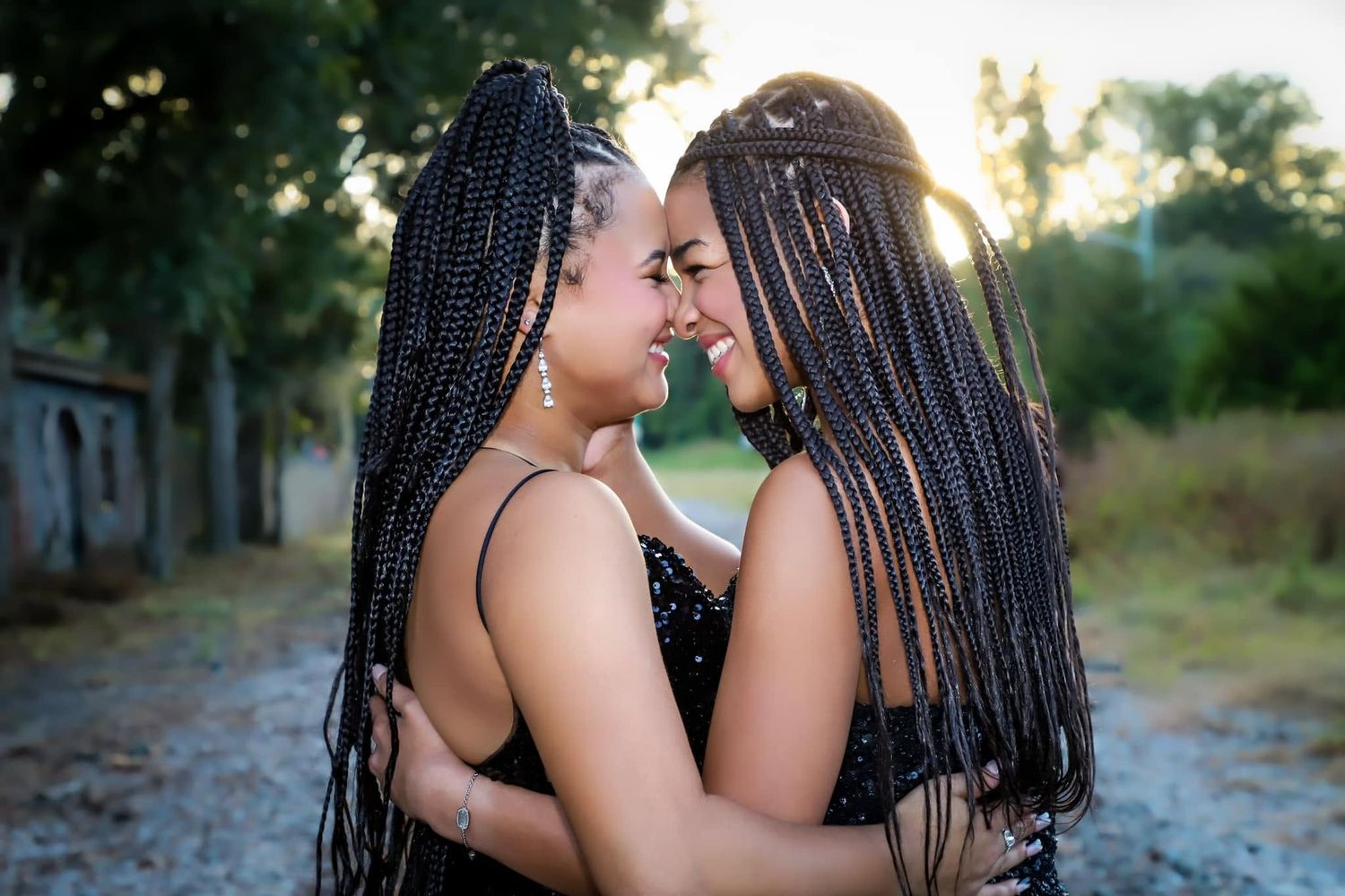 Two young women with long box braids embracing and smiling face to face outdoors at sunset.