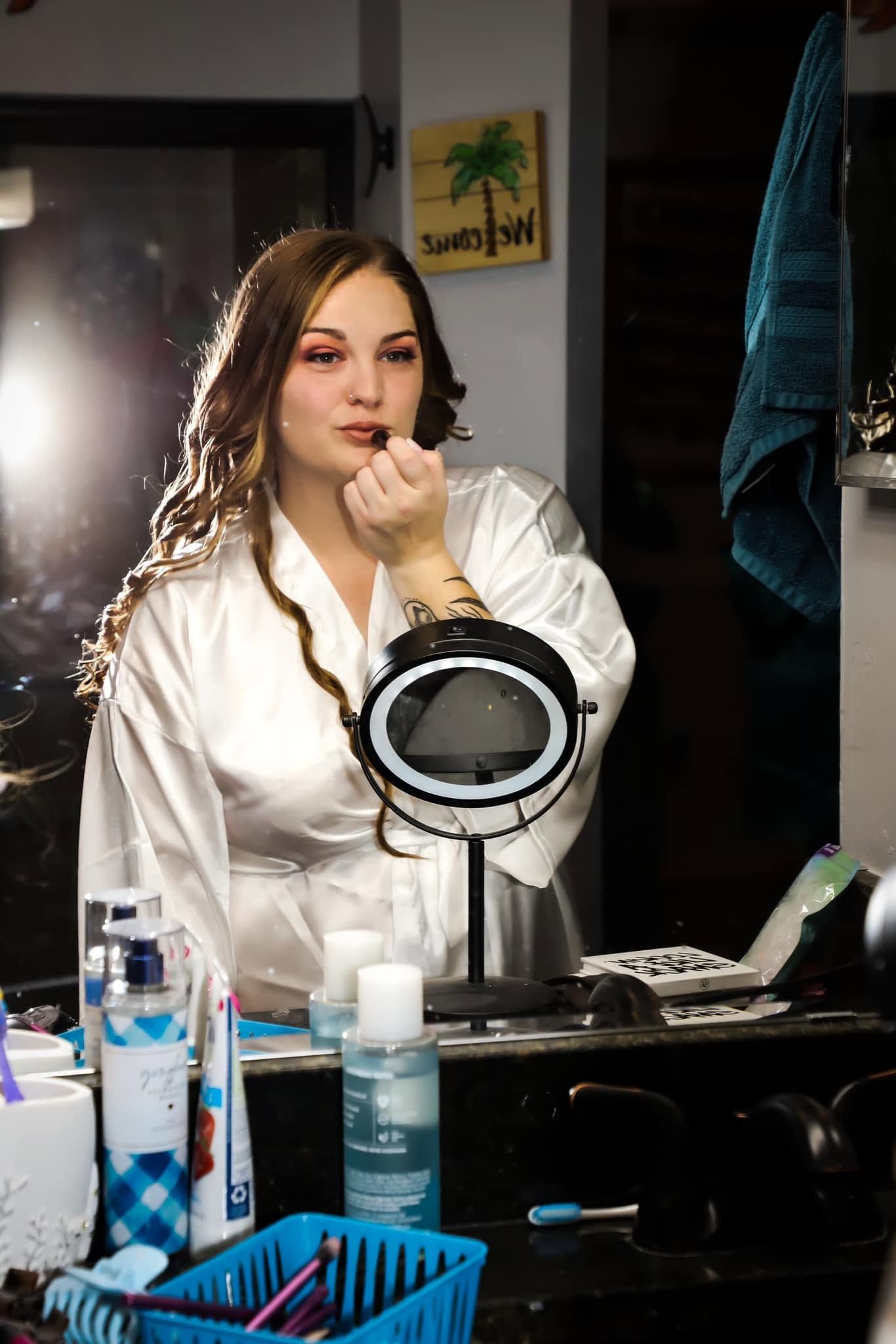 Bride in a white satin robe applies lipstick at a vanity while looking in a mirror before her wedding in Gainesville, Florida.