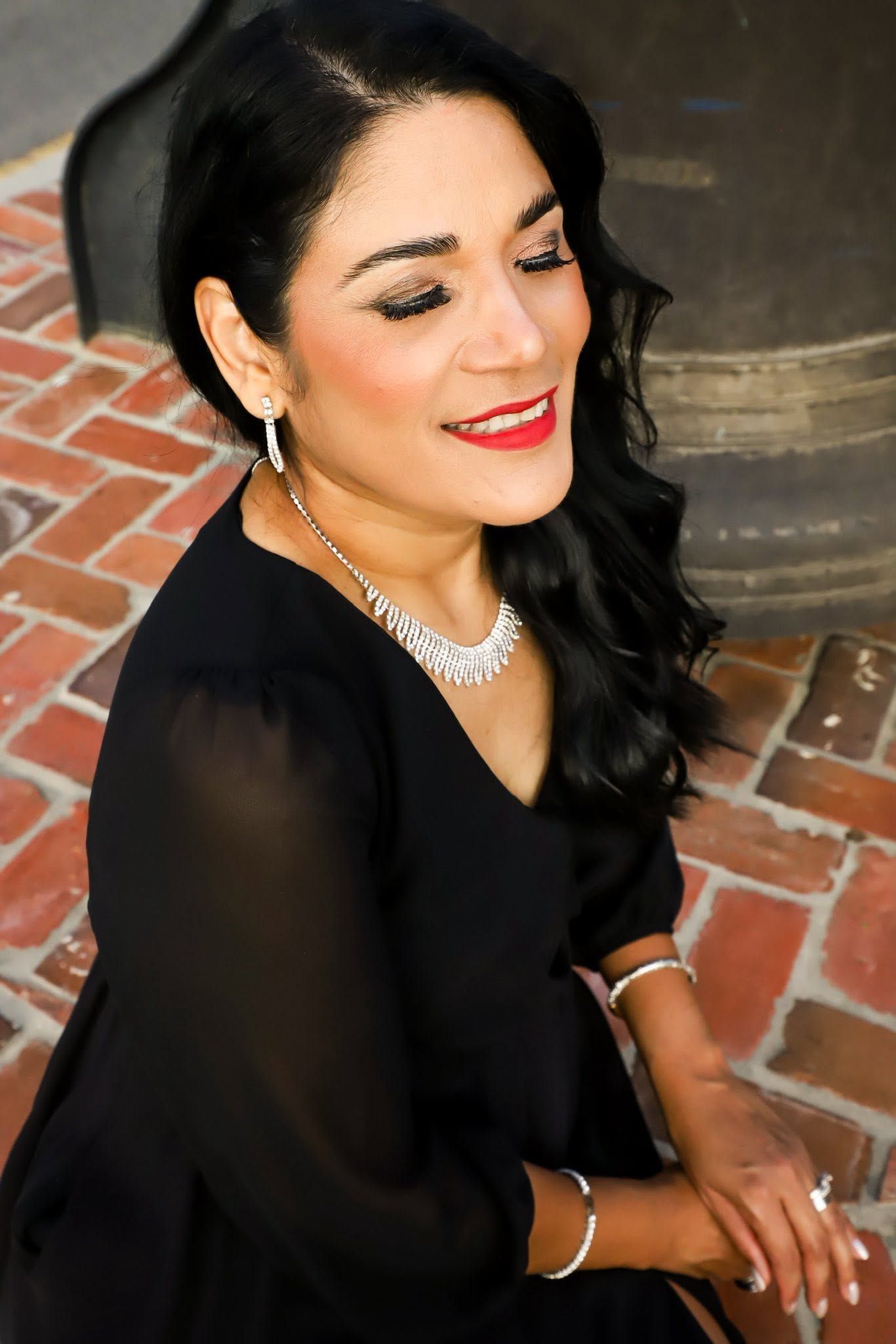 Elegant outdoor portrait of a woman in a black dress and diamond necklace, smiling softly against a brick background in Ocala, Florida.