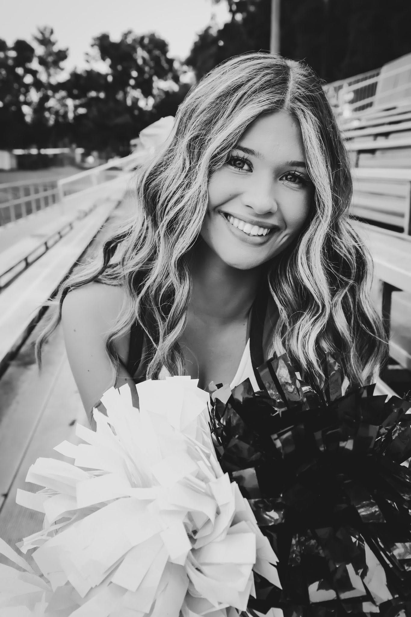 Black and white senior portrait of a smiling high school cheerleader holding pom poms in stadium bleachers in Gainesville, Florida.
