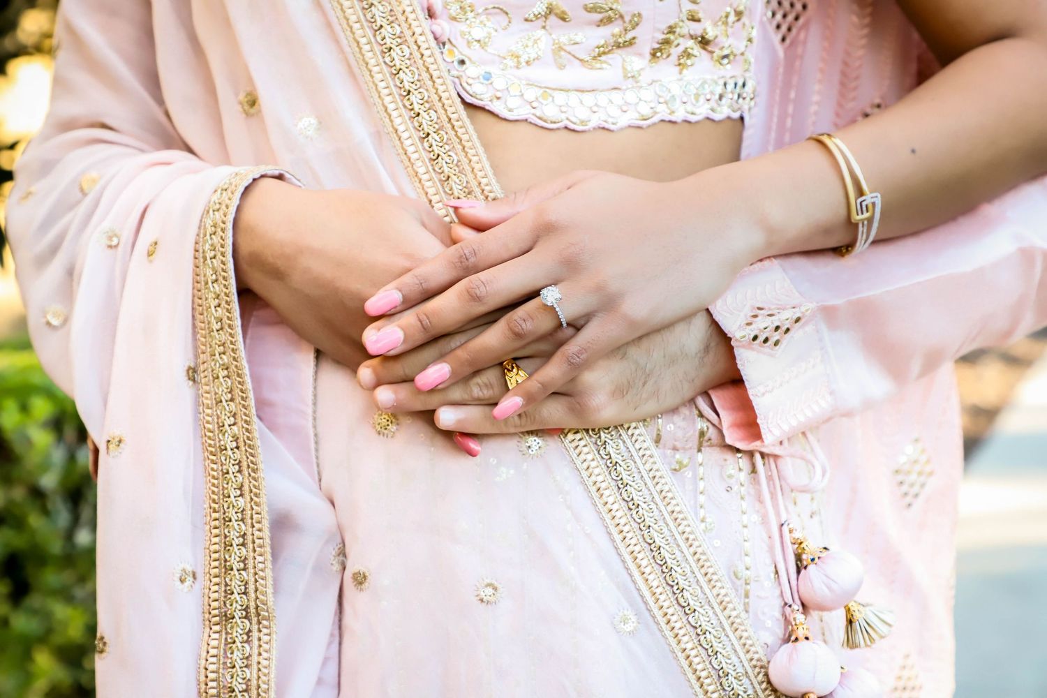 A close-up of a couple holding hands, showcasing an engagement ring and gold bangles in pink attire.