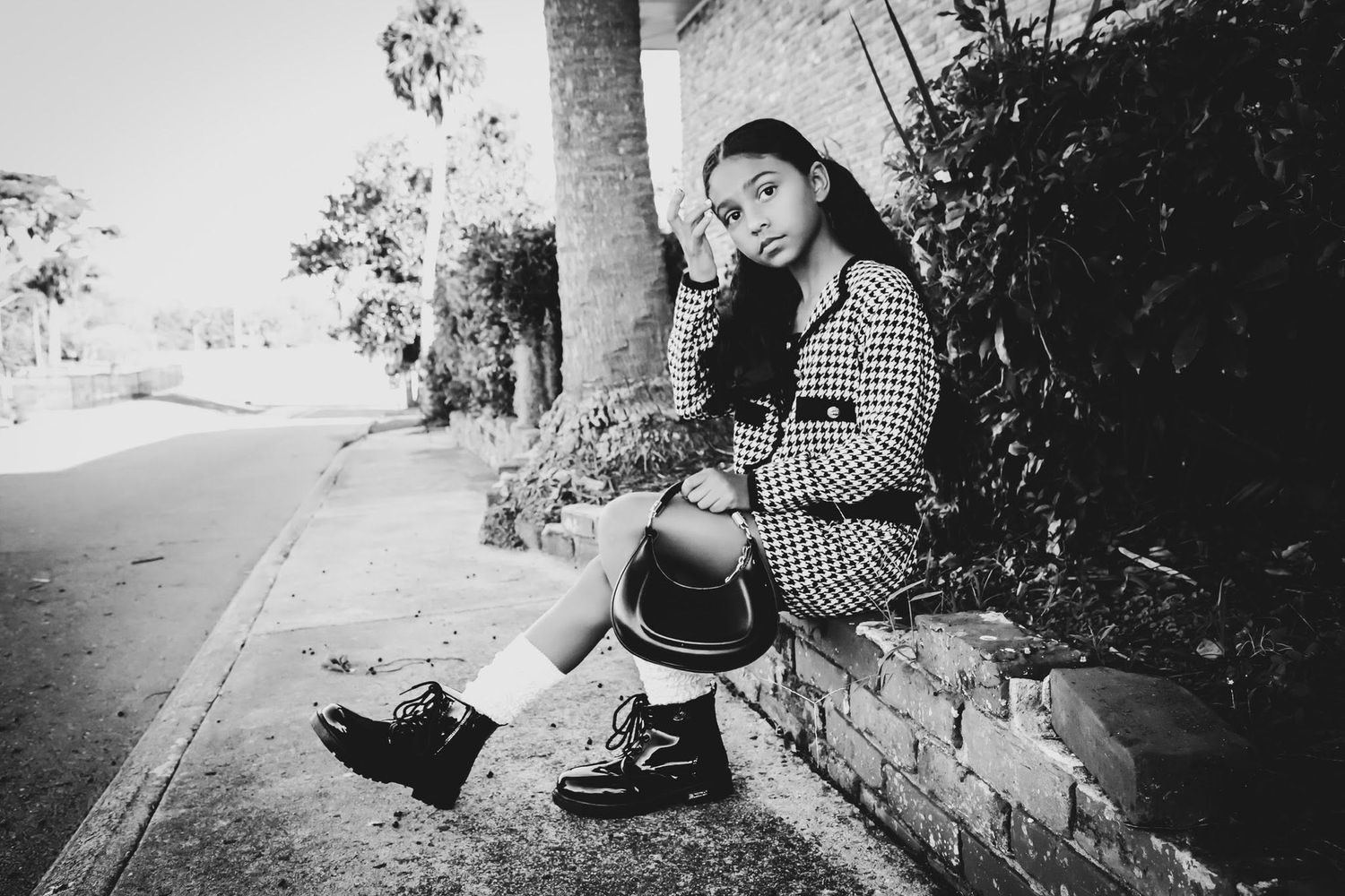 Black and white portrait of a young girl sitting on a curb beside a brick wall in a checkered outfit.