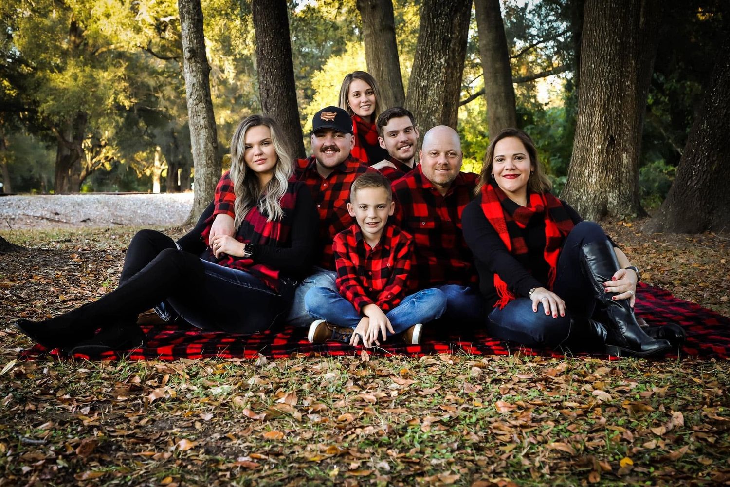 A large family in red plaid outfits sits together on fallen autumn leaves in a wooded park.