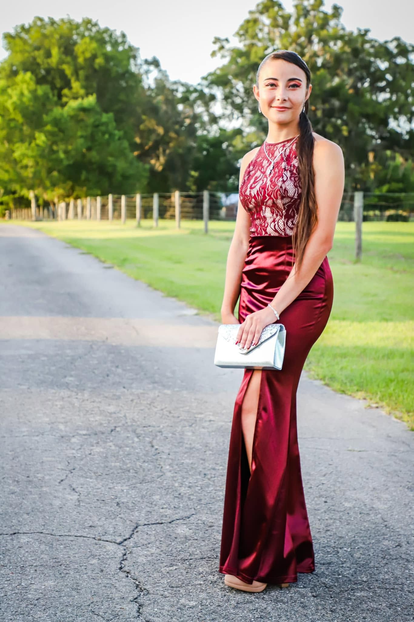 Prom portrait of a teen in a burgundy satin gown holding a silver clutch on a rural road at sunset in Newberry, Florida.