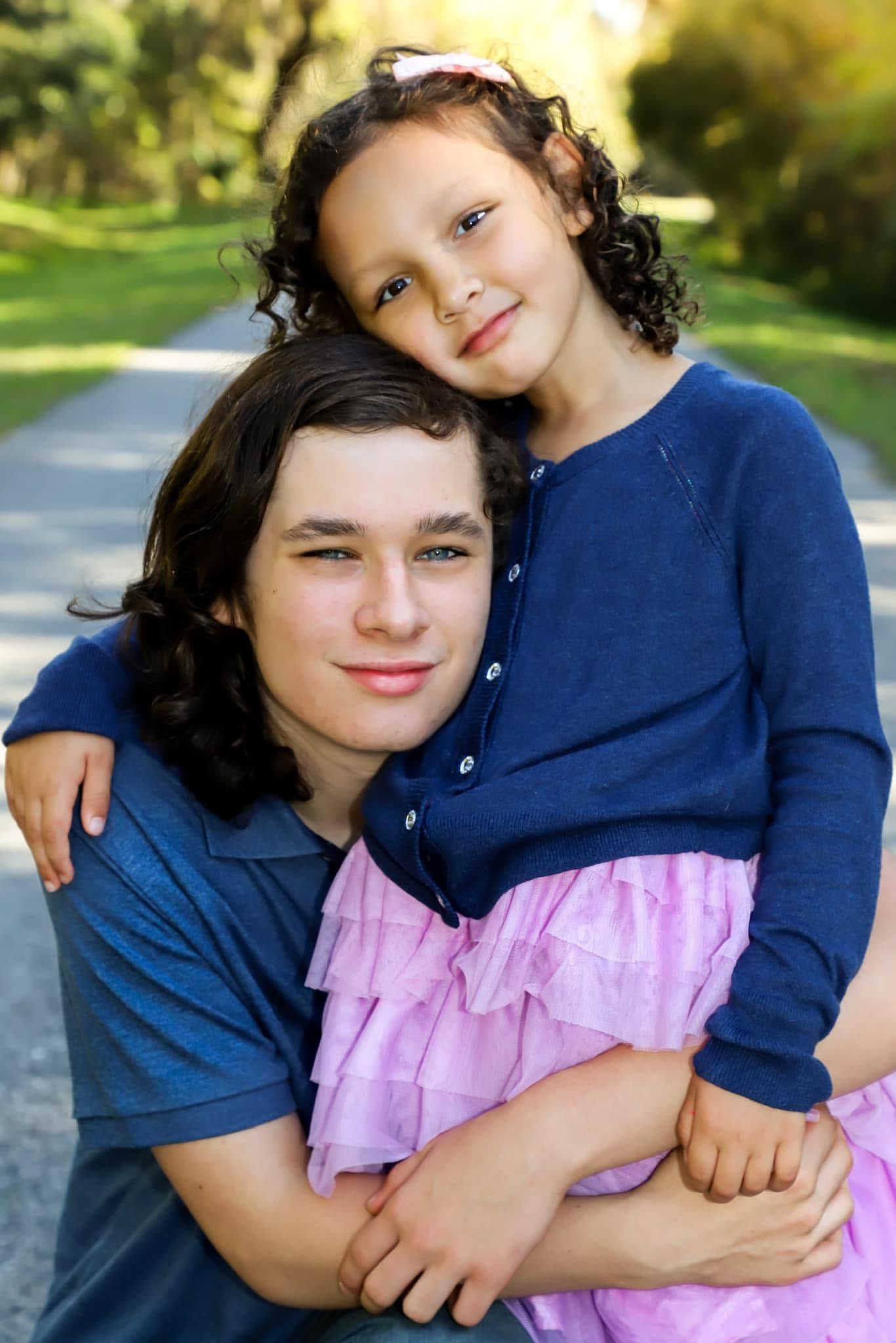 Family portrait of a young man and curly-haired girl in a pink dress sharing a warm embrace on a park path in Lake City, Florida.