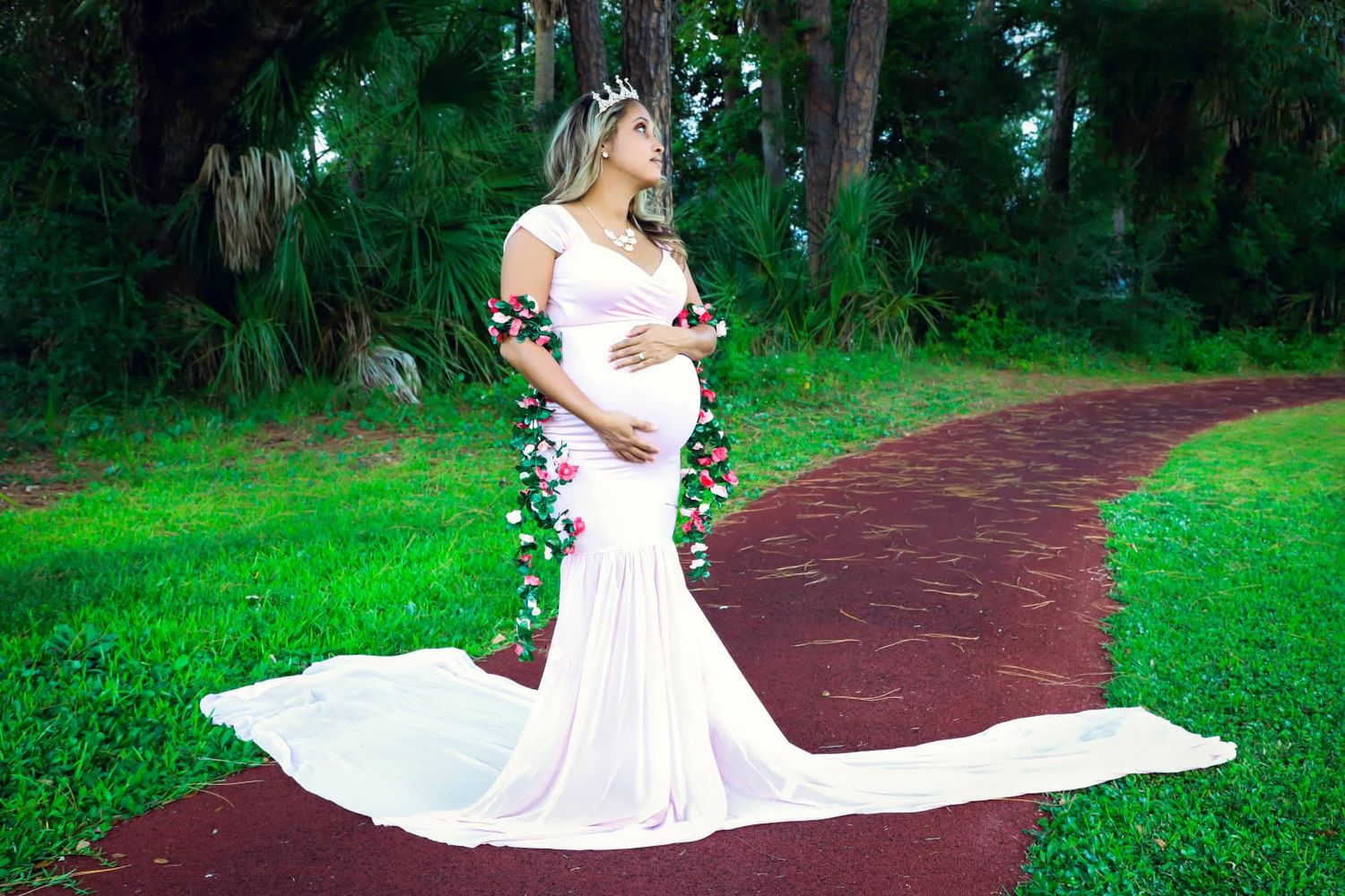 Elegant pregnant woman in white floral mermaid maternity gown posing on red brick garden path surrounded by greenery in savannah Georgia.