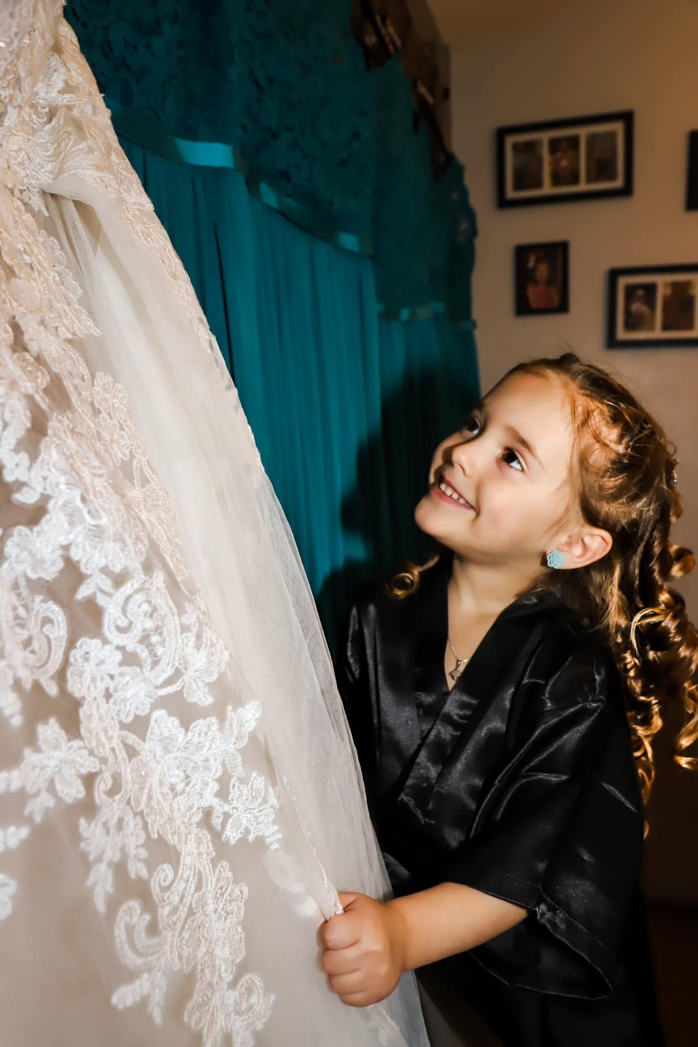 A curly-haired little girl in a black robe smiles while touching an elegant lace wedding dress on a hanger.