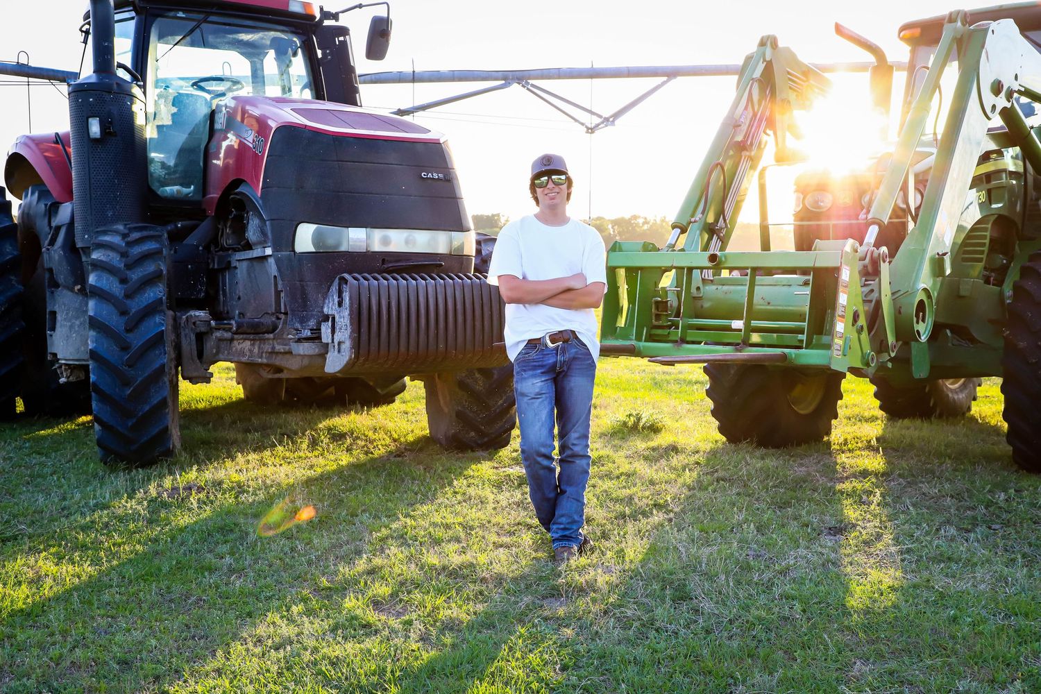 A farmer in jeans and a cap stands confidently between large agricultural tractors at golden hour.