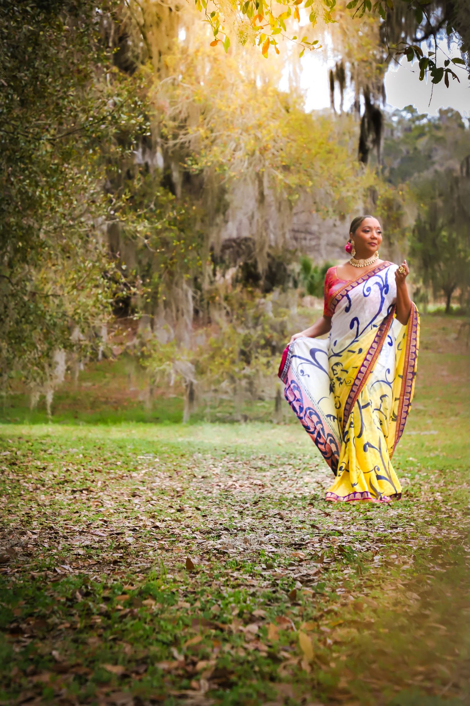 Woman in colorful traditional sari walking gracefully under Spanish moss-draped trees outdoors.