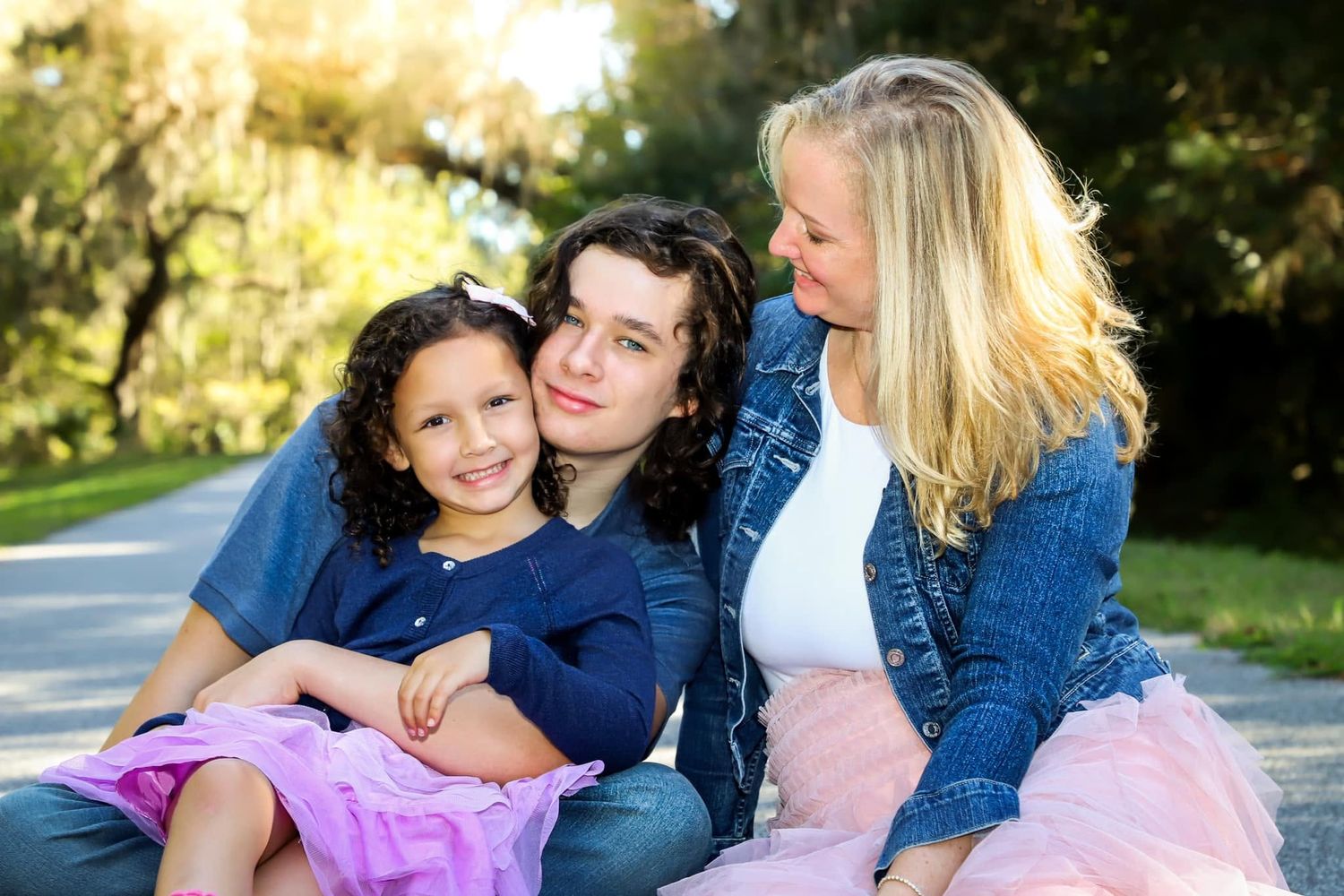 Two siblings hugging a young girl in a pink tutu skirt outdoors in a park.
