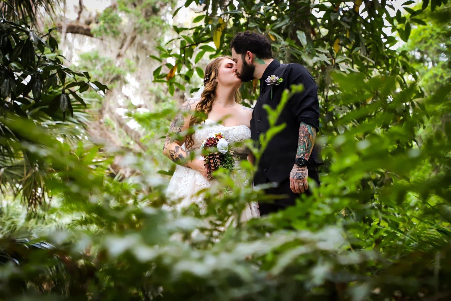 Bride and groom share a romantic kiss surrounded by lush green foliage in an outdoor garden setting.