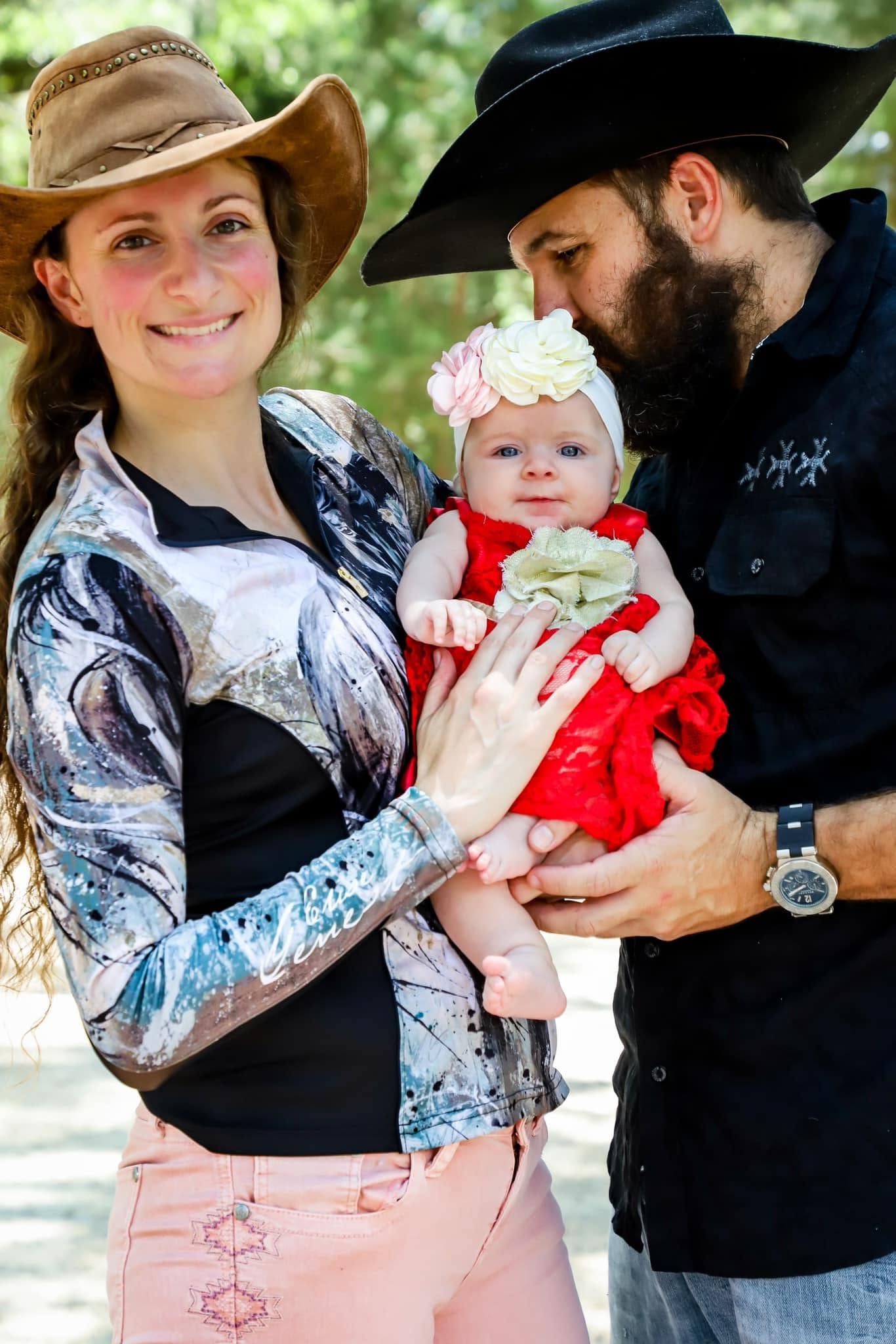 Family and Horse Photography in Starke Florida. Smiling couple in cowboy hats holding adorable baby in red dress outdoors in dappled light.