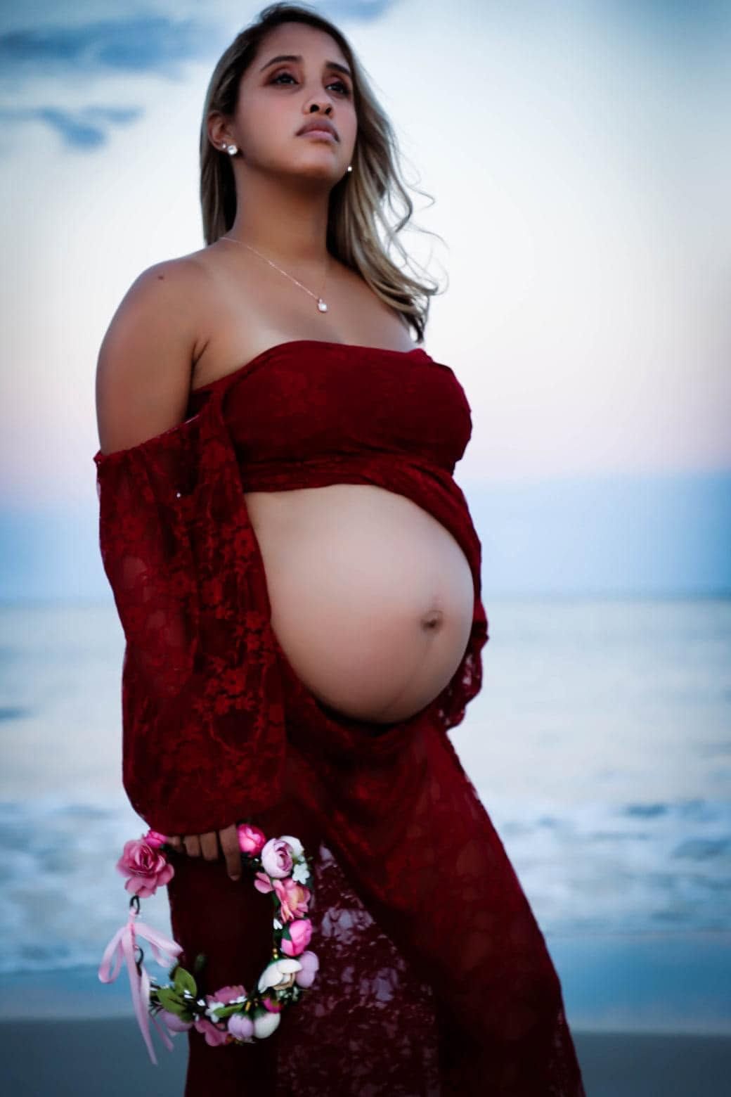 A pregnant woman in a red lace gown holds a floral wreath on a beach at sunset in Panama City Beach