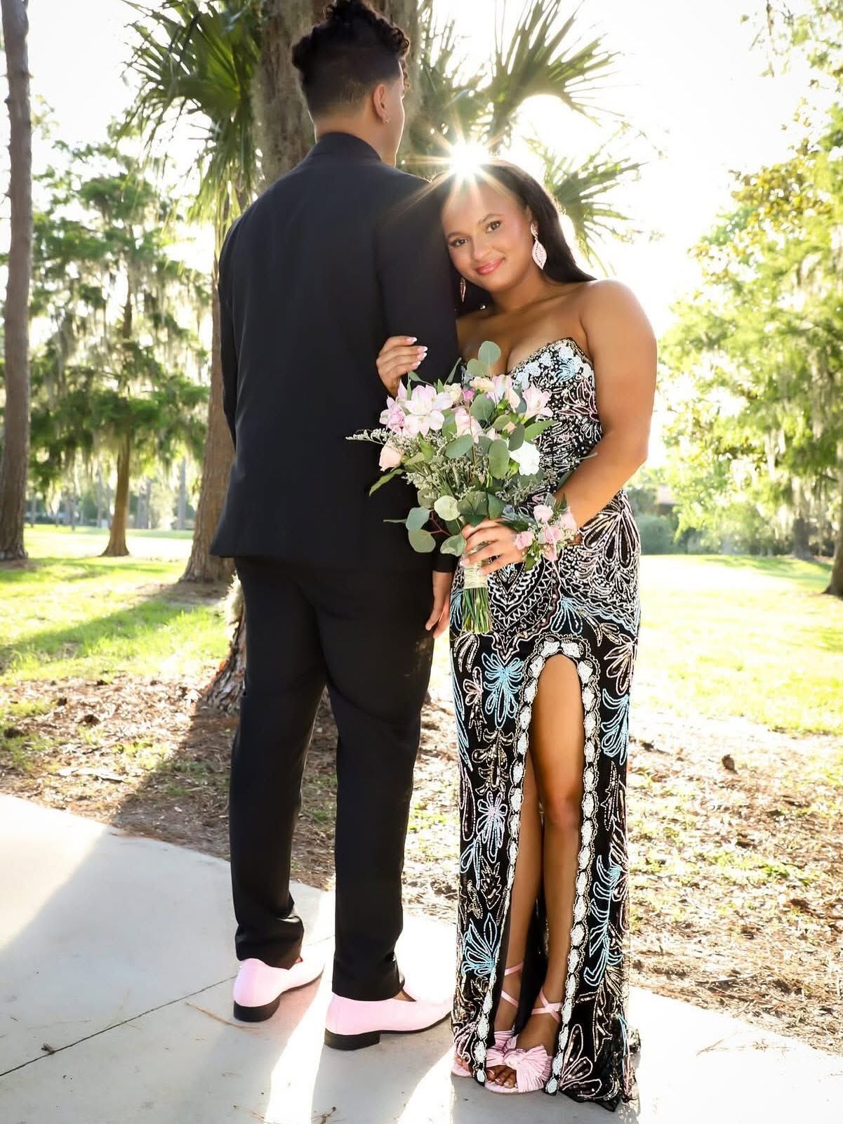Couple in formal attire posing with a bouquet of flowers in a sunlit park.