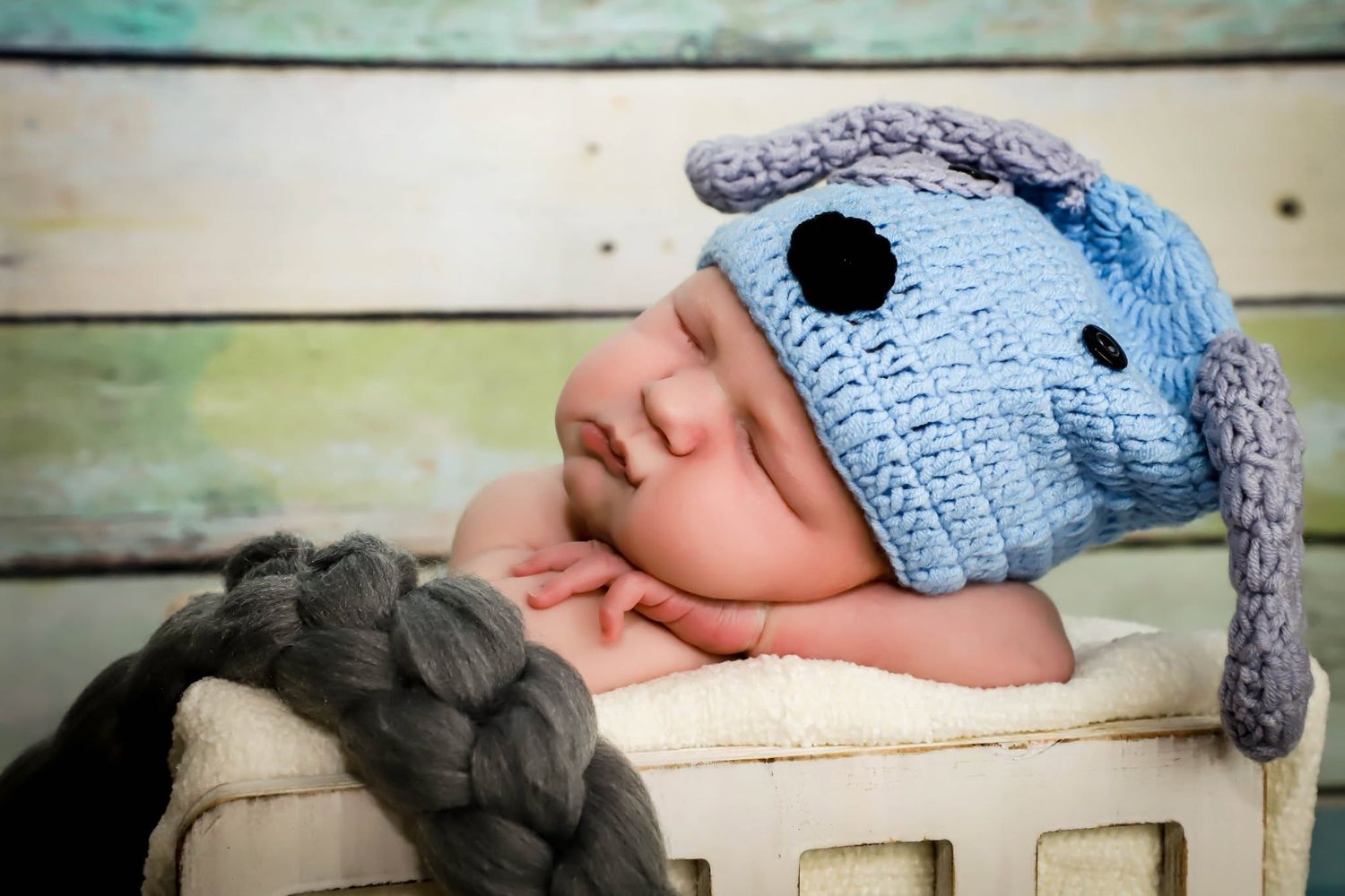 Sleeping newborn baby wearing blue crocheted puppy hat in wooden basket.