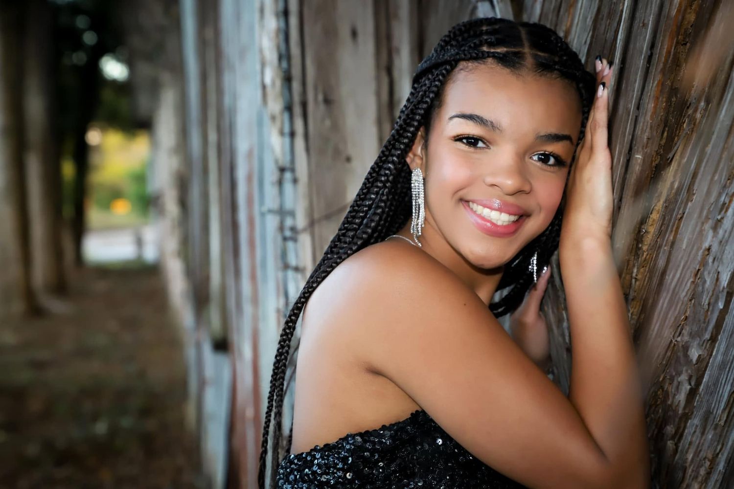 A smiling young woman with long braids leans against a rustic wooden wall in a sparkling top.