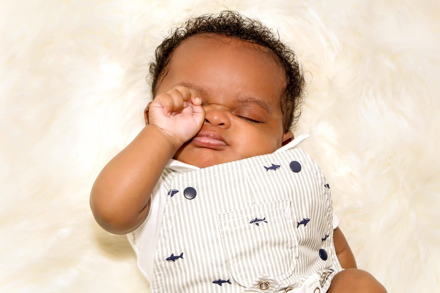 Adorable sleeping newborn baby in white striped onesie with shark print, resting on fluffy cream background.