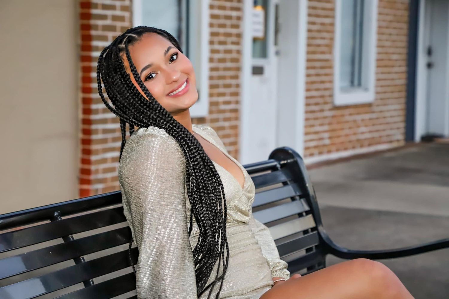 Young woman with long box braids smiling on a bench outside a brick building.