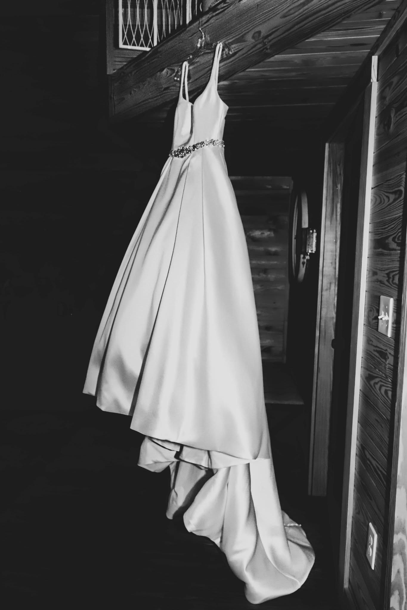 Elegant white A-line wedding gown with embellished waistband hanging on wooden staircase in black and white photo.