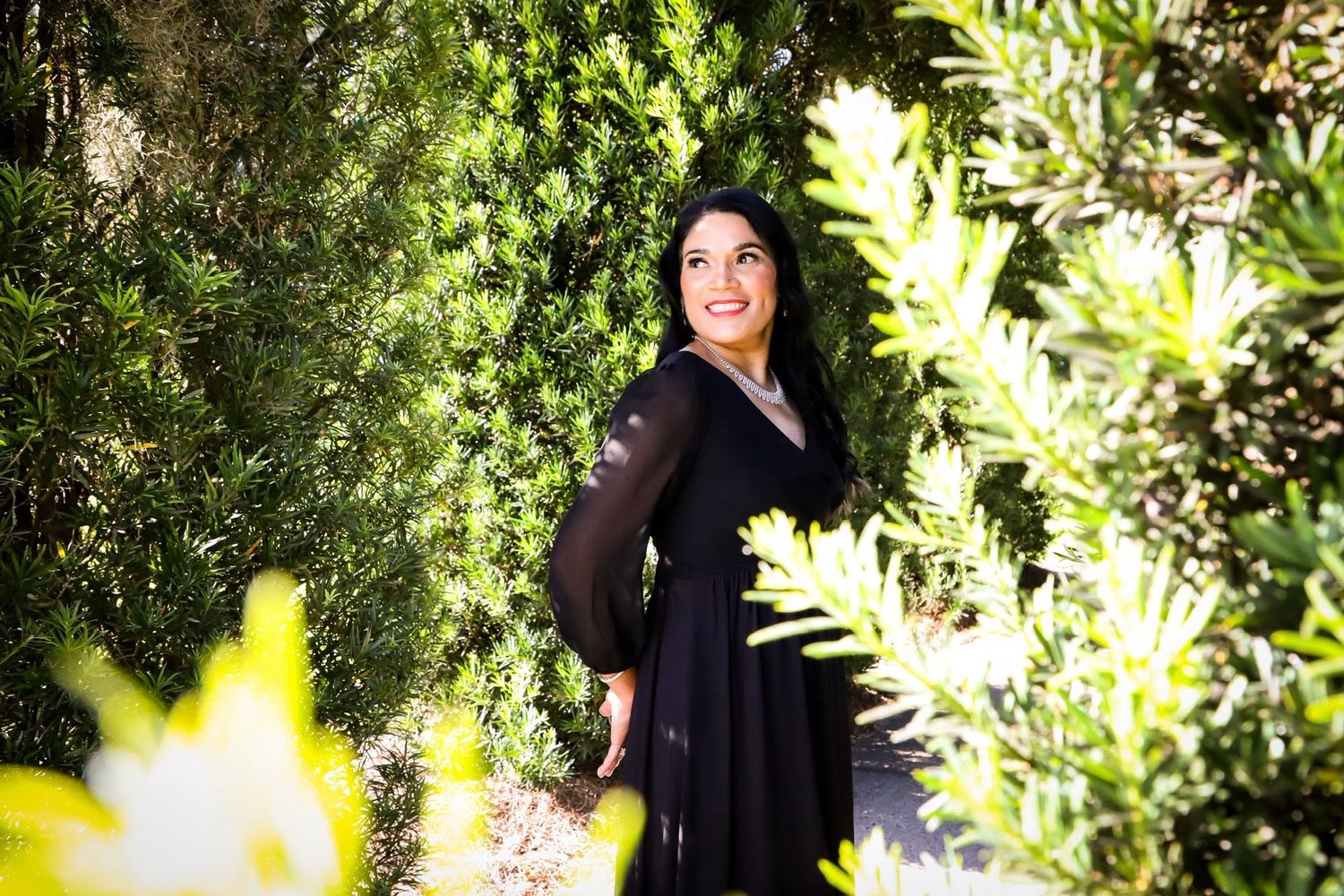 Outdoor portrait of a smiling woman in a black dress standing among lush green bushes in bright sunlight in Ocala, Florida.