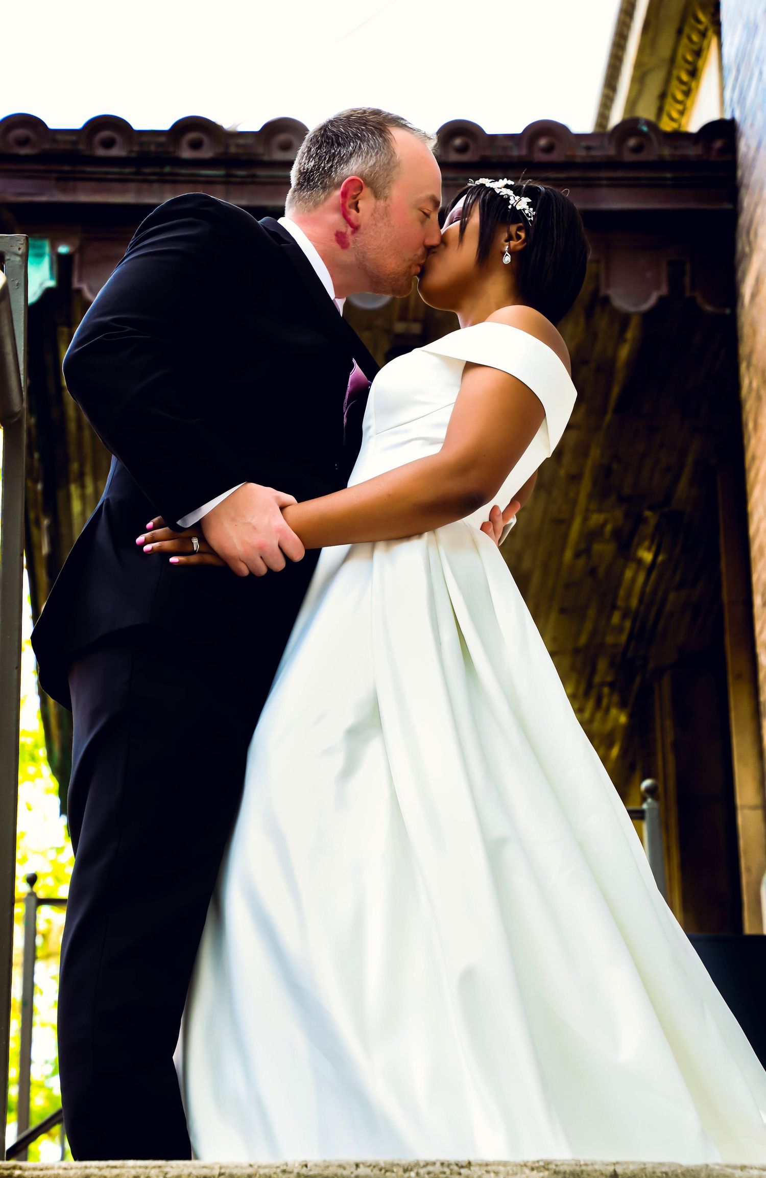 Interracial couple sharing first kiss as newlyweds, groom in black tuxedo and bride in elegant white off-shoulder ball gown.