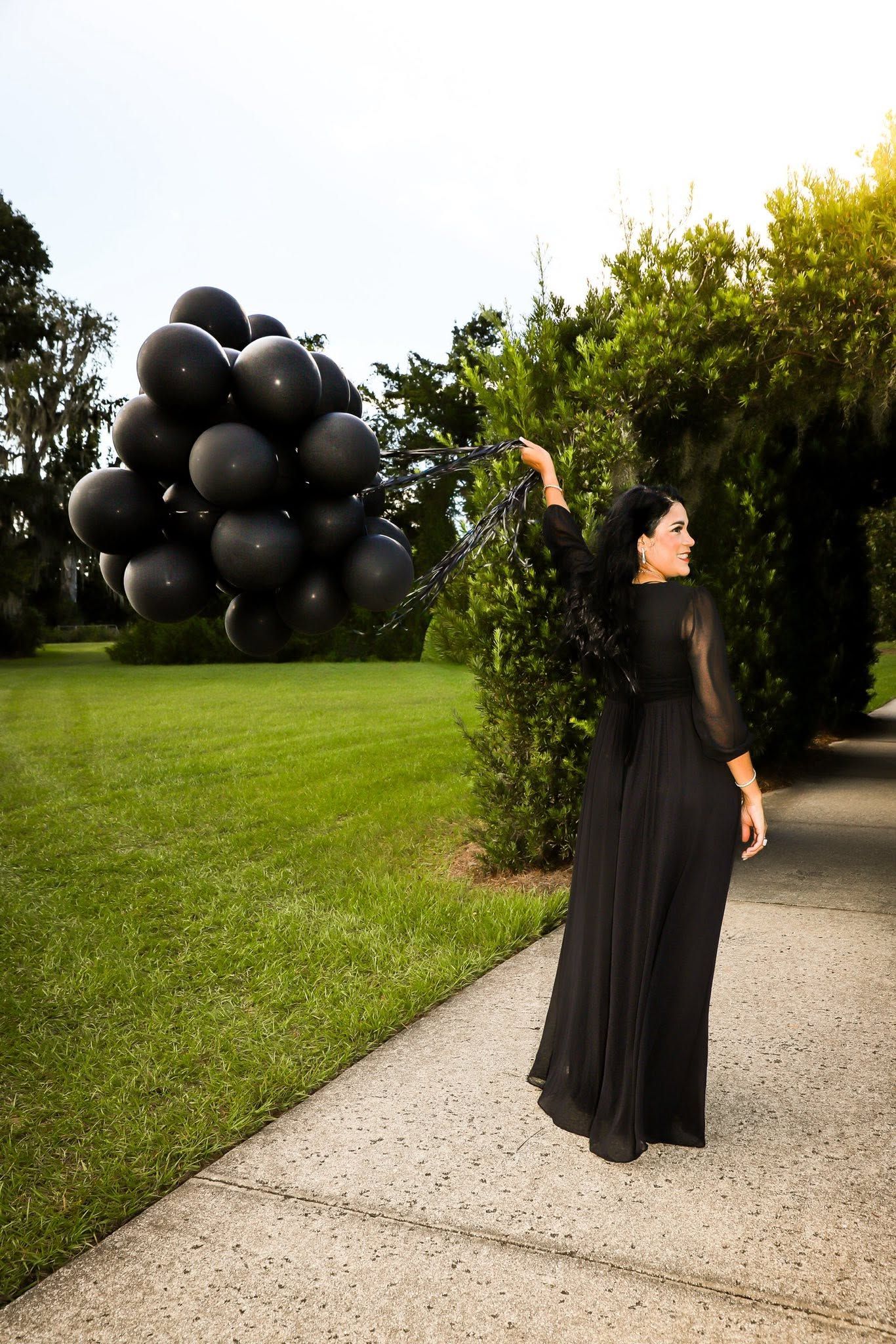 50th Birthday photo session with woman in black gown holding a bunch of black balloons on a garden pathway.