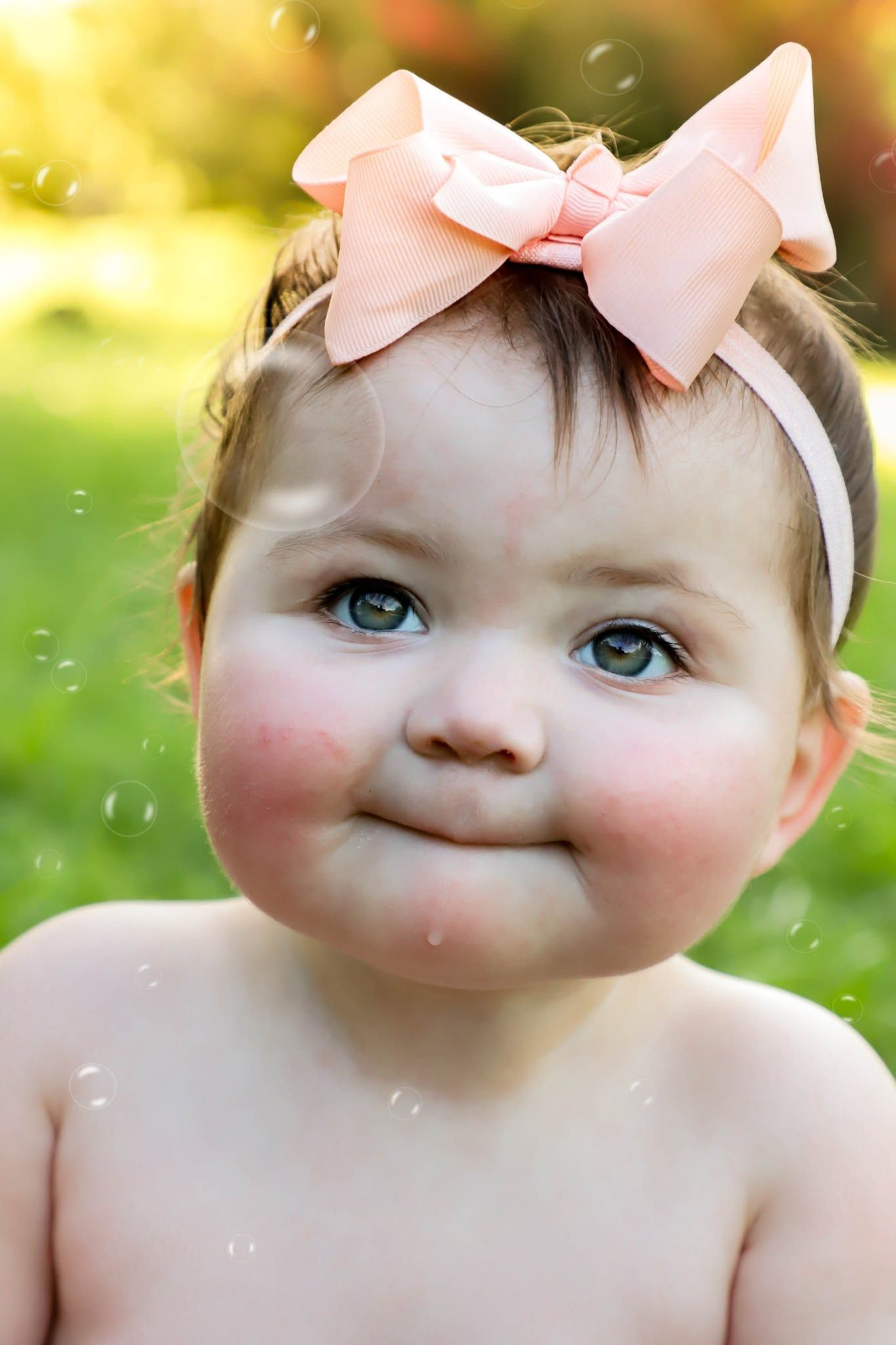 Adorable baby girl with pink bow headband smiling outdoors with green bokeh background.