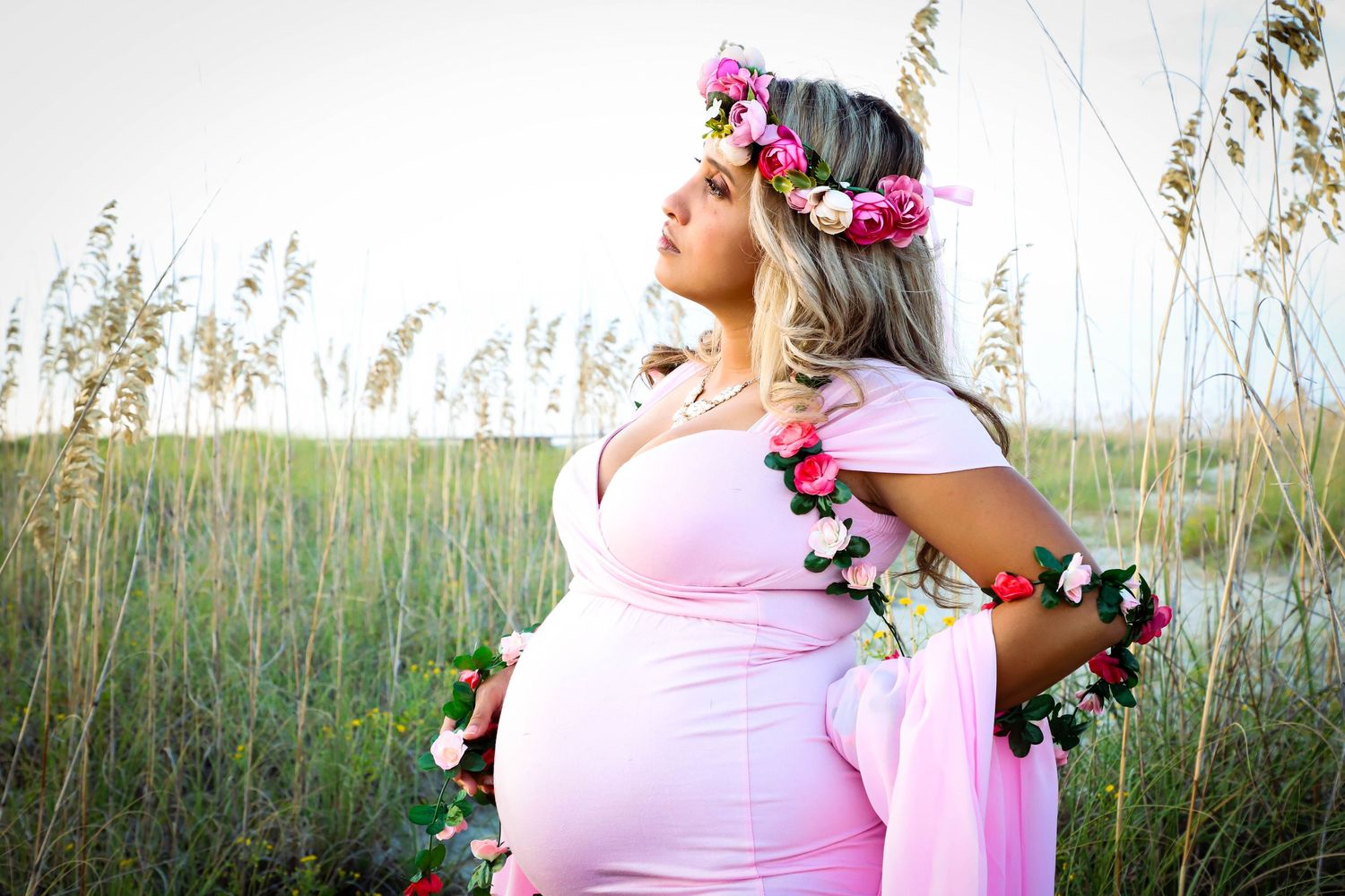 A pregnant woman in a pink gown wears a floral crown while posing gracefully in a field of tall grass.