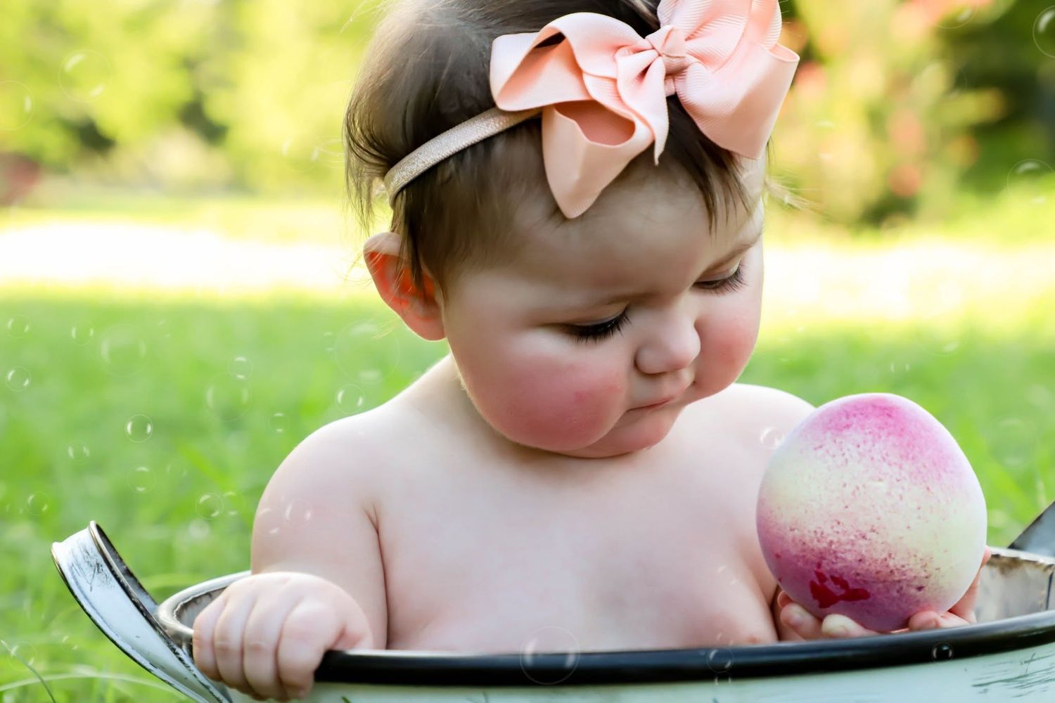 Baby girl with a pink bow headband sitting in a tub holding a peach outdoors in Gainesville, Florida.