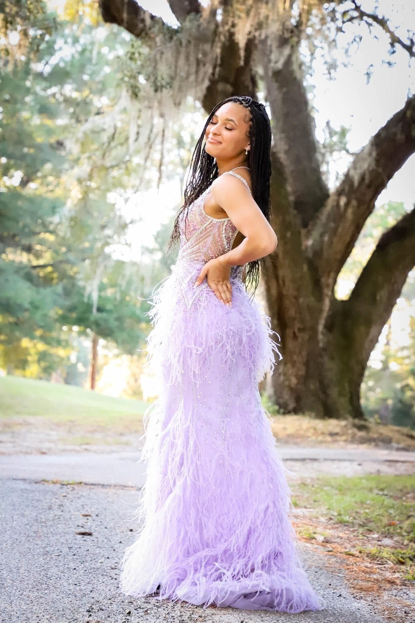 Young woman in lavender feathered gown posing gracefully beside a large oak tree.