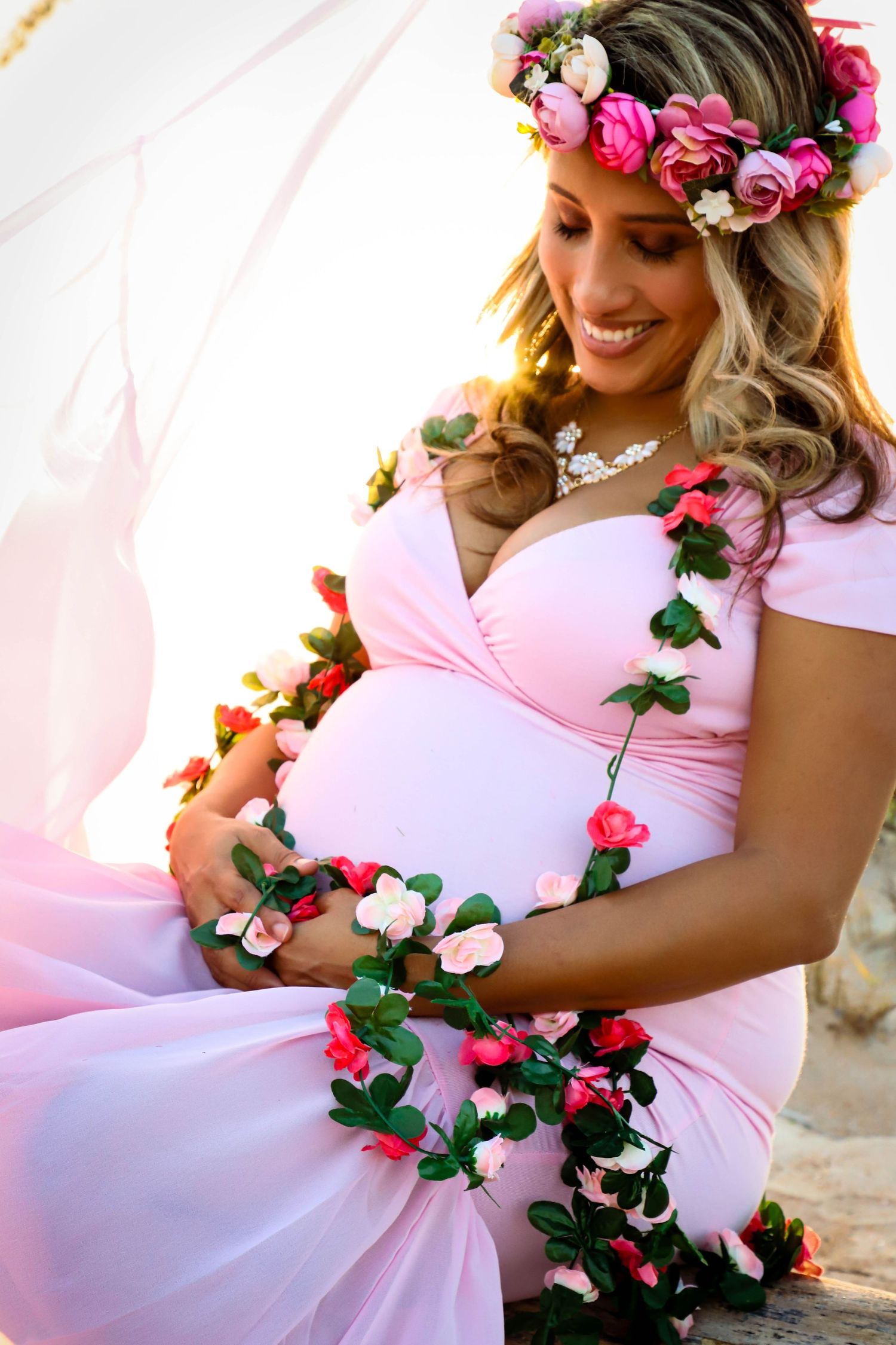 Pregnant woman in pink floral gown wearing a flower crown cradling her baby bump outdoors.