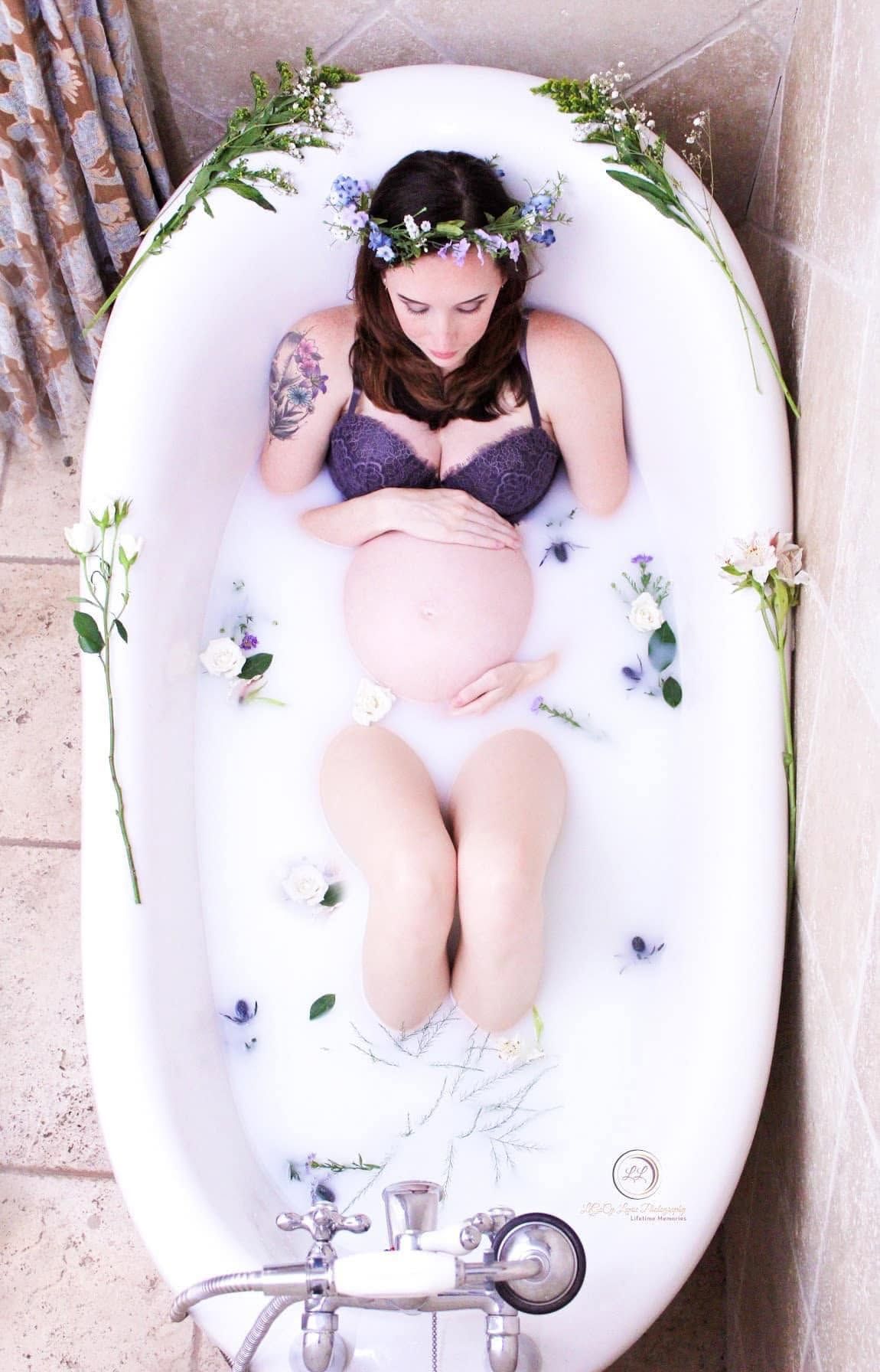 Pregnant woman in floral crown relaxing in milk bath with flowers and greenery.
